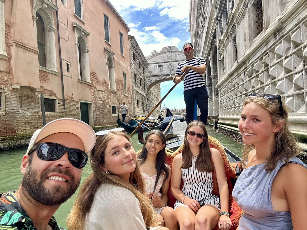 Teen travelers ride a gondola under the Bridge of Sighs in Venice as a gondolier in a striped shirt rows behind them