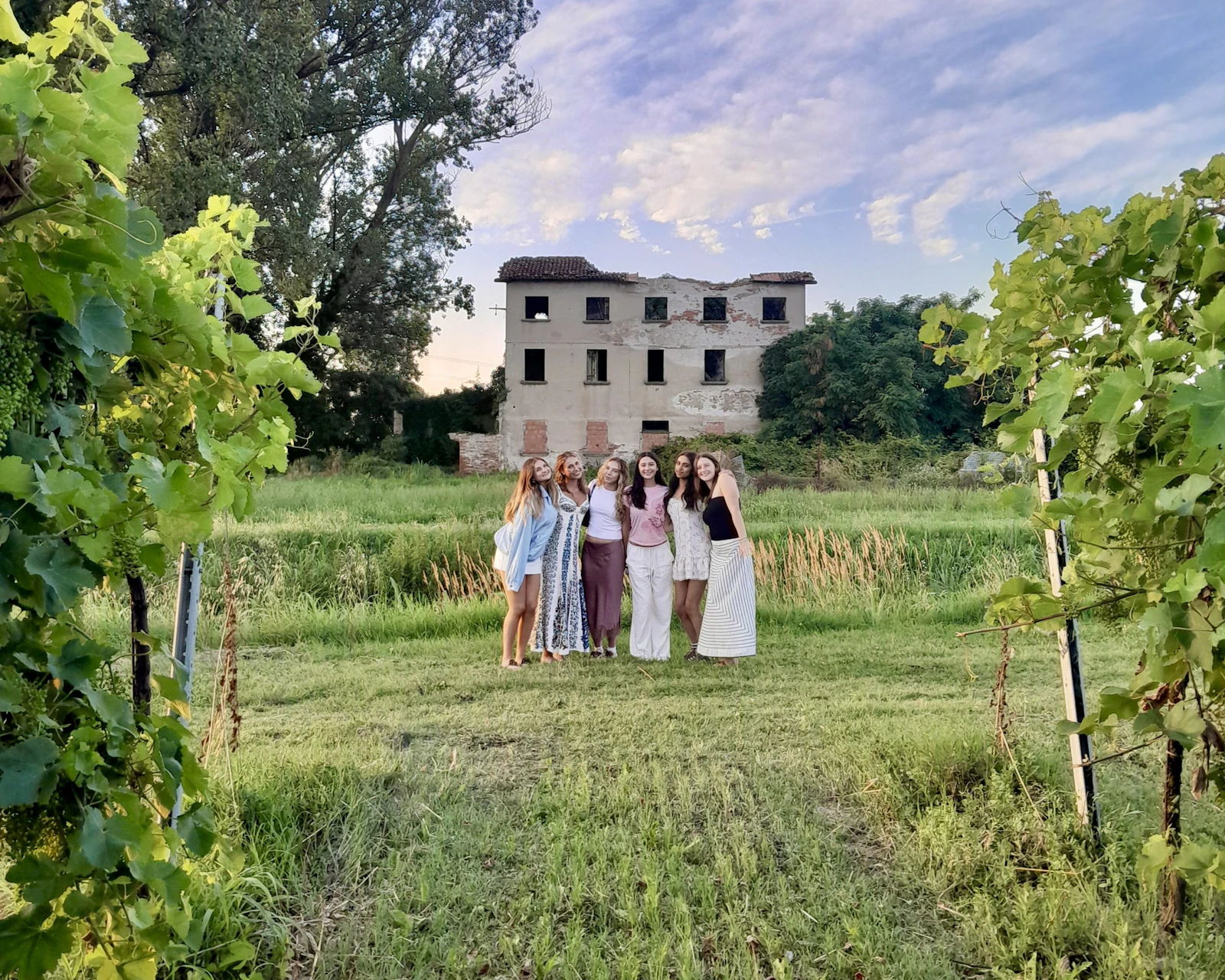 Six teen travelers stand together in a mown clearing at dusk in Italy, framed by grapevines, with an old villa behind