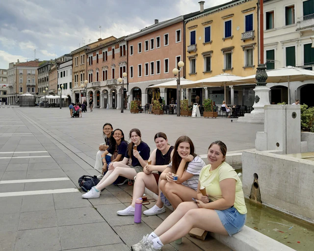 Six teen travelers sit on the rim of a stone fountain in a wide Italian piazza lined with pastel arcades