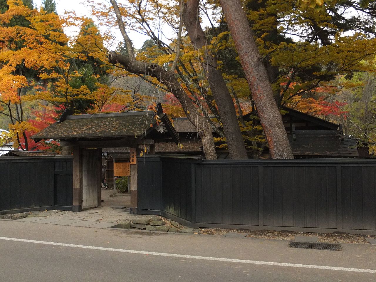 A wooden gate and fence surround a traditional samurai house beneath autumn foliage