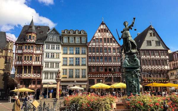 A row of steep-gabled, half-timbered townhouses on a cobbled square, with a green bronze figure of Justice holding scales above a fountain in the foreground.