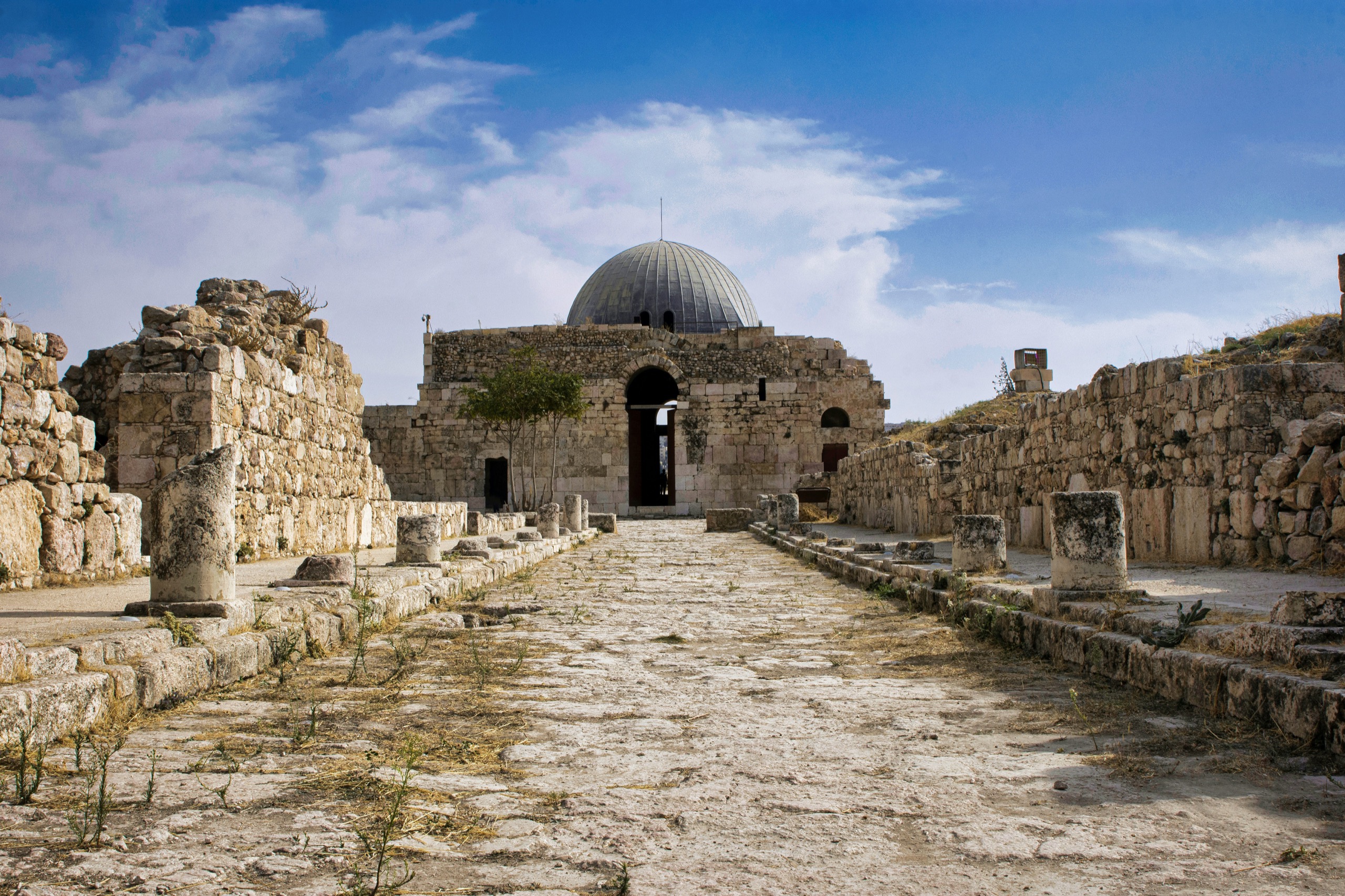 A domed stone hall stands at the end of a ruined colonnaded street on a hilltop.