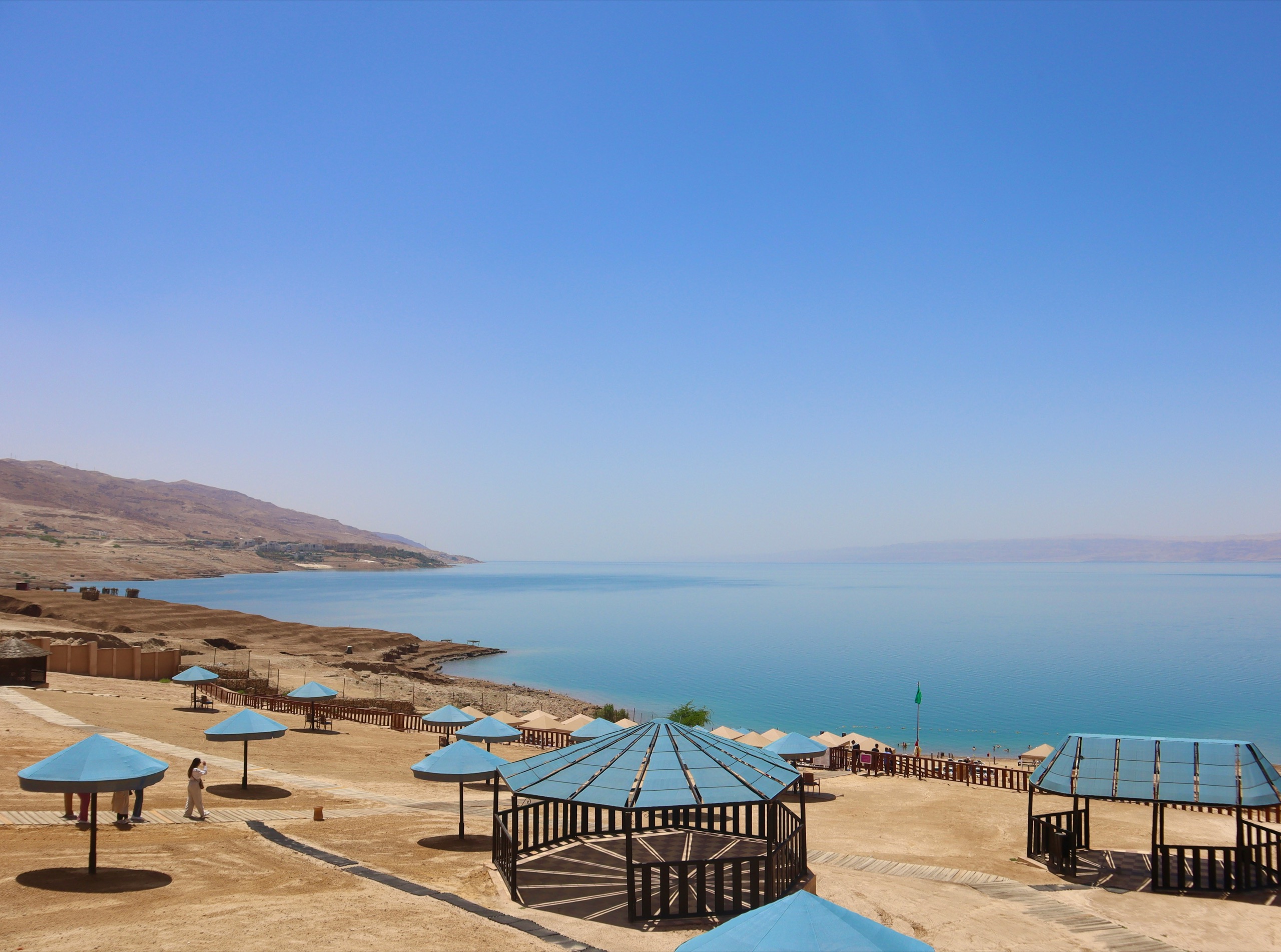 Blue shade canopies stand on a stony shore beside calm pale water, with hills across the bay.