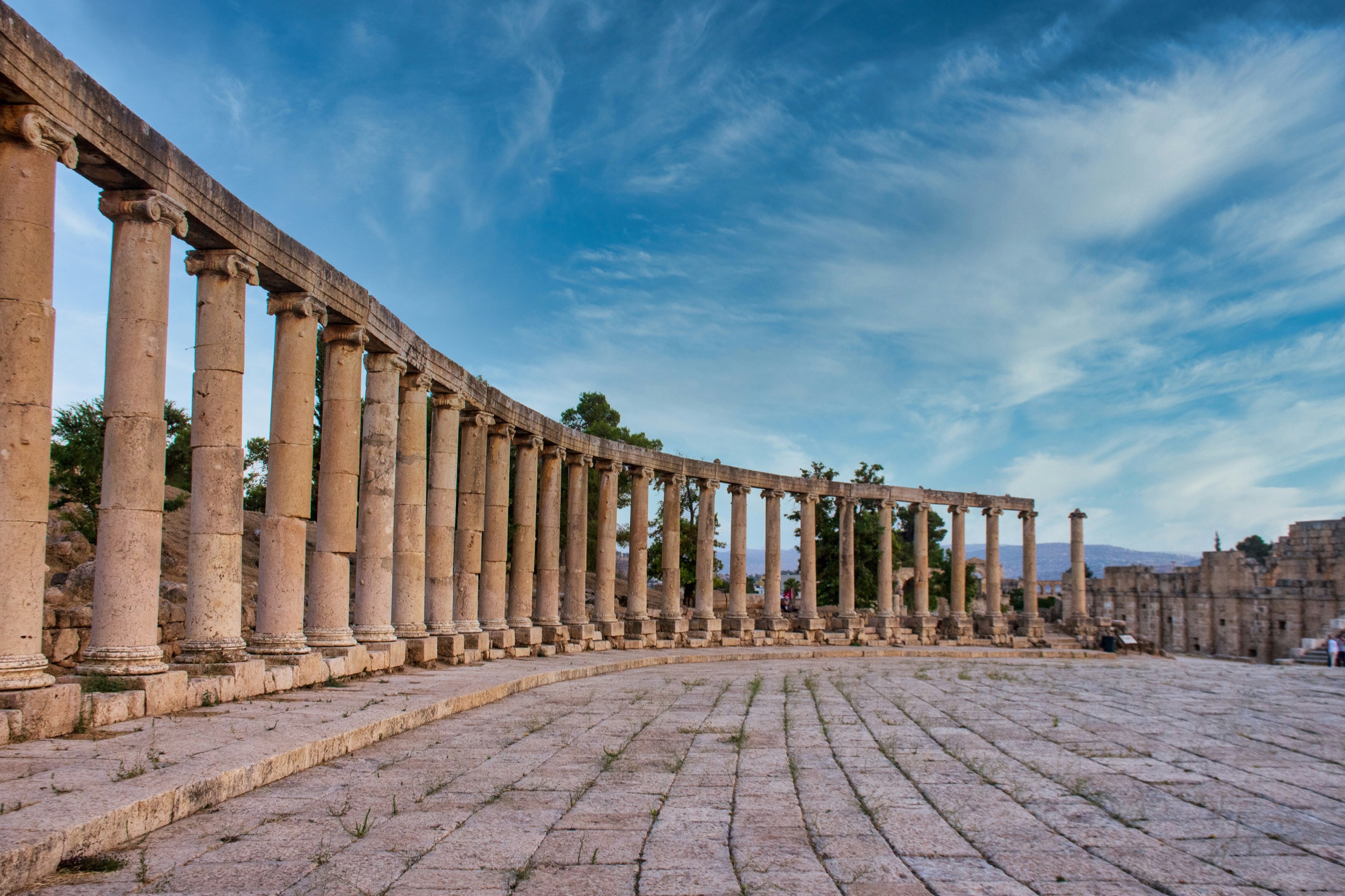 A curved colonnade of stone columns encloses a paved oval plaza under wispy cloud.