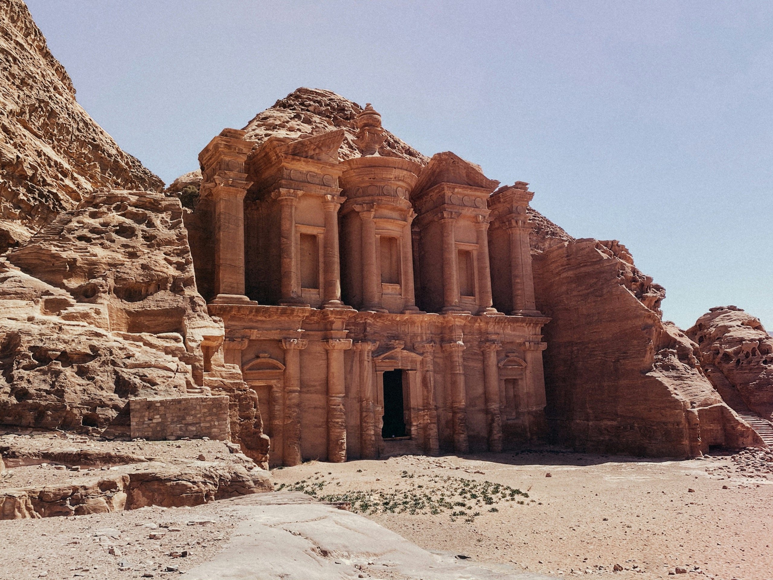 A wide rock-cut facade with columns and an urn stands above a bare sandy terrace.