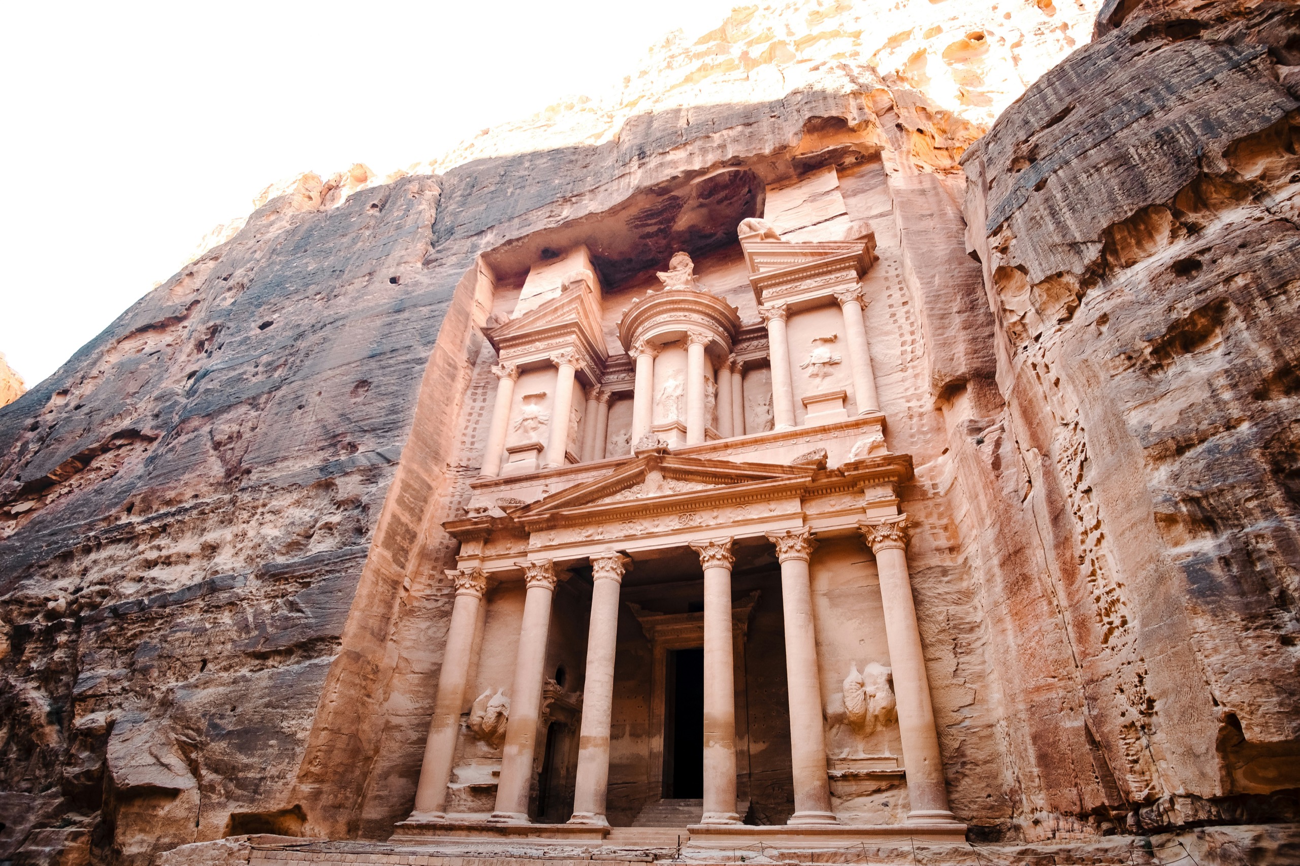 A rose-colored facade cut into the rock, a wedge of sky above its columns and small figures at its base.