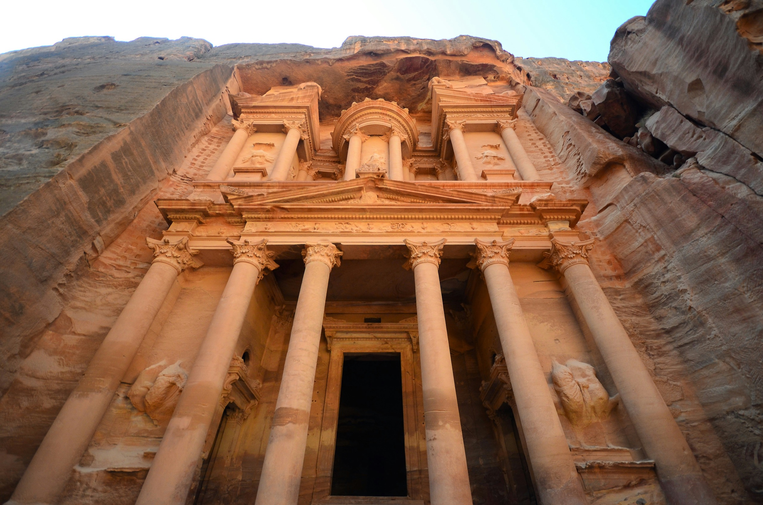 A carved classical facade rises from the cliff face, seen from below against a strip of sky.