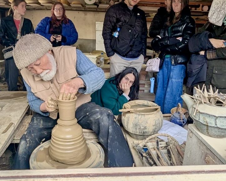 A potter throws a tall vessel on the wheel while students watch from behind the bench