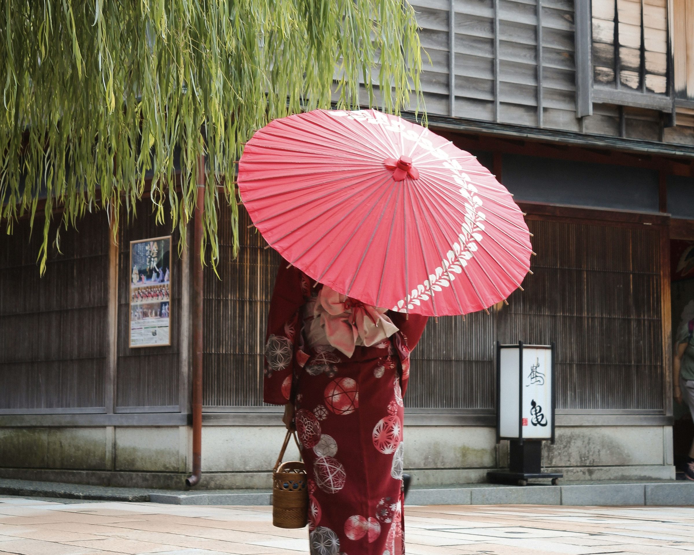 A woman in a red kimono carries a paper parasol past wooden teahouse fronts