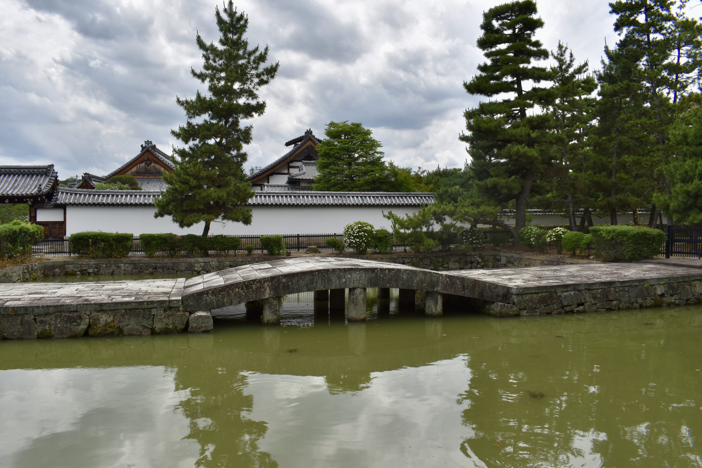 A stone arch bridge crosses a temple moat toward a white plaster wall and tiled roofs