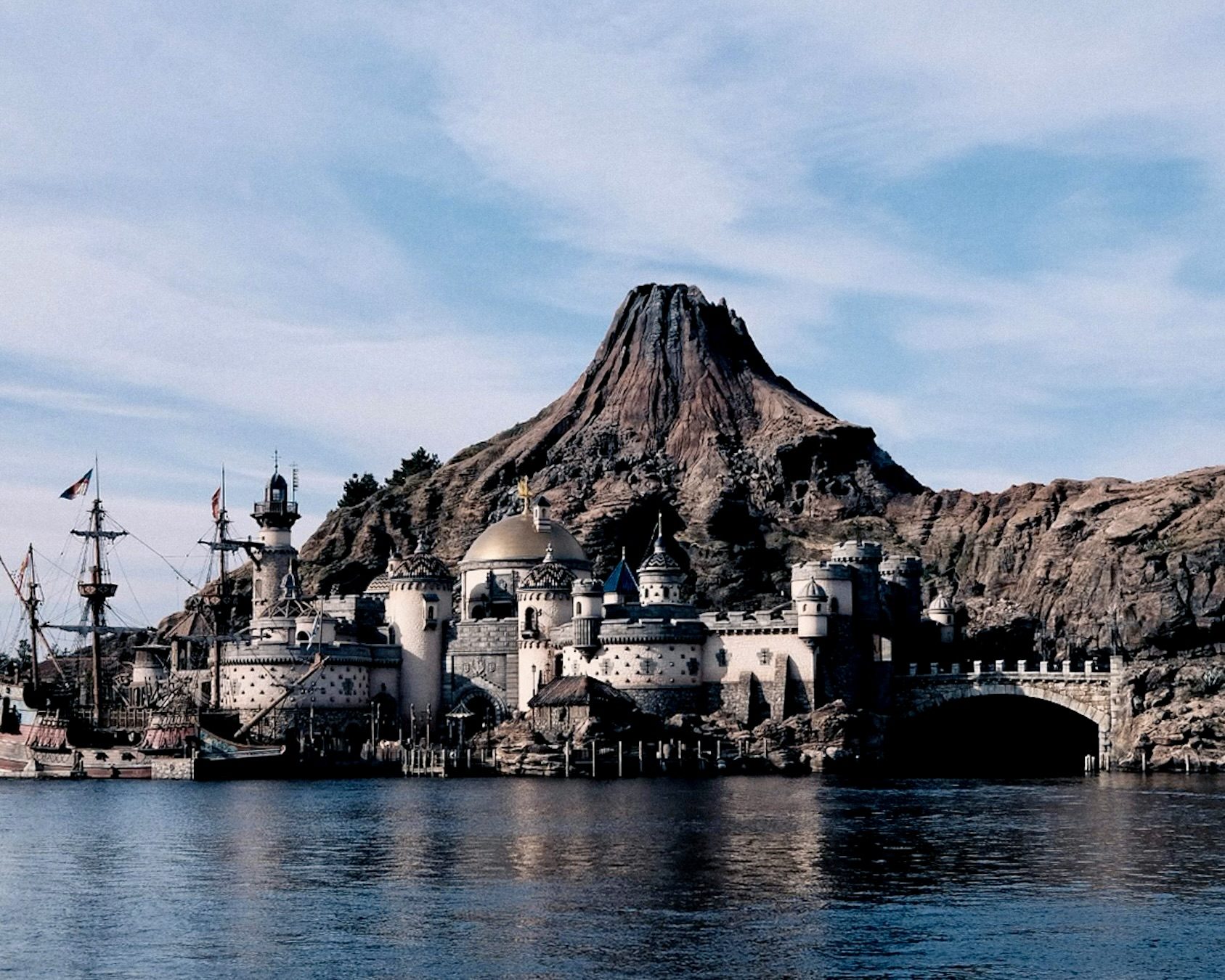 A volcano peak rises behind a harbour fortress and a tall ship at Tokyo DisneySea and closing dinner