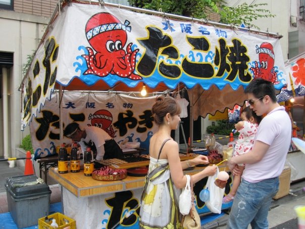 A family stands in front of a Takoyaki stall in a food market.