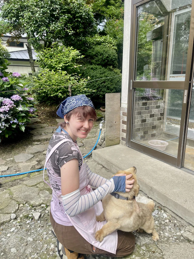 A smiling teen traveler in a blue bandana and pink apron kneels on a stone path and pets a tan puppy beside a house.
