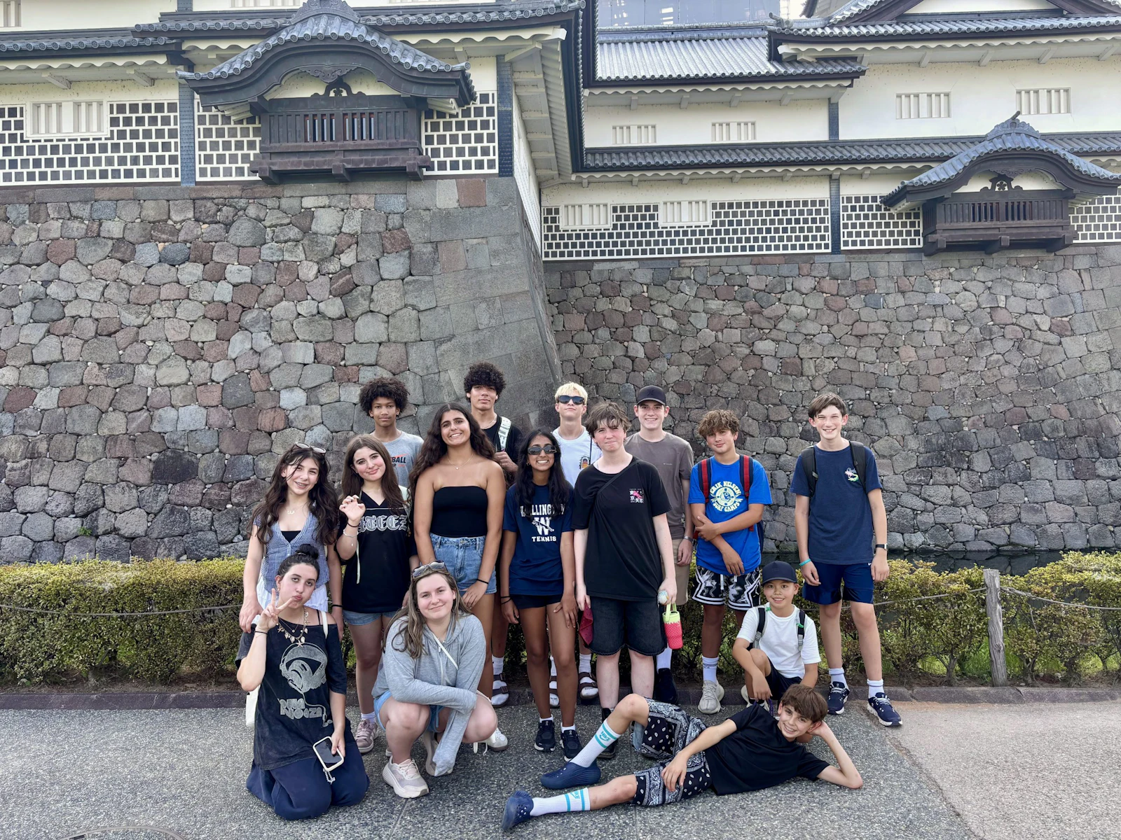 A group of teen travelers poses on a pavement in front of a tall stone castle wall topped with a white turreted gatehouse.