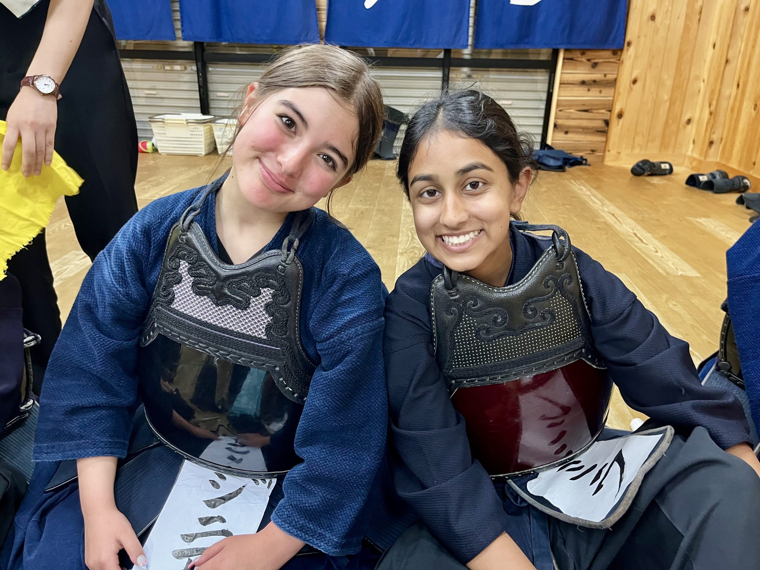 Two teen travelers in indigo kendo armor sit side by side on a dojo floor, smiling at the camera.
