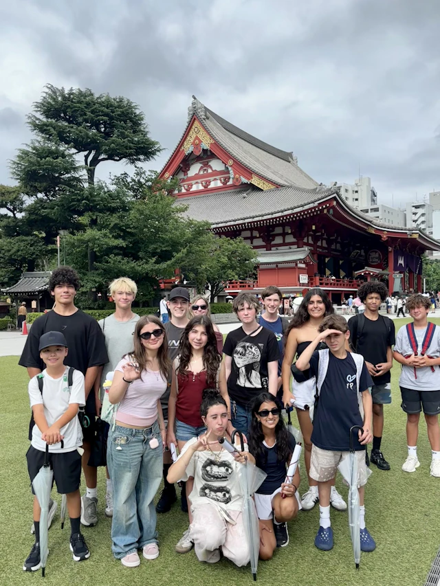 A group of teen travelers poses on a lawn in front of a red-roofed Japanese temple hall and a pine tree under a grey sky.