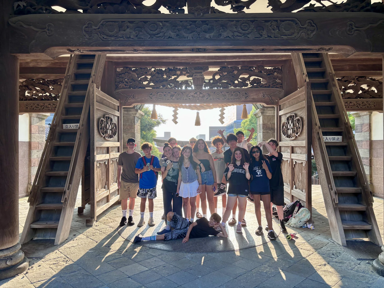 Teen travelers stand together in a carved wooden temple gateway, backlit by afternoon sun, with stairs rising on both sides.