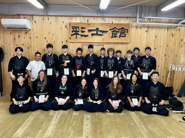 Teen travelers in dark kendo uniforms kneel in rows on a wooden dojo floor beneath a calligraphy sign, with a few in street clothes at the front.