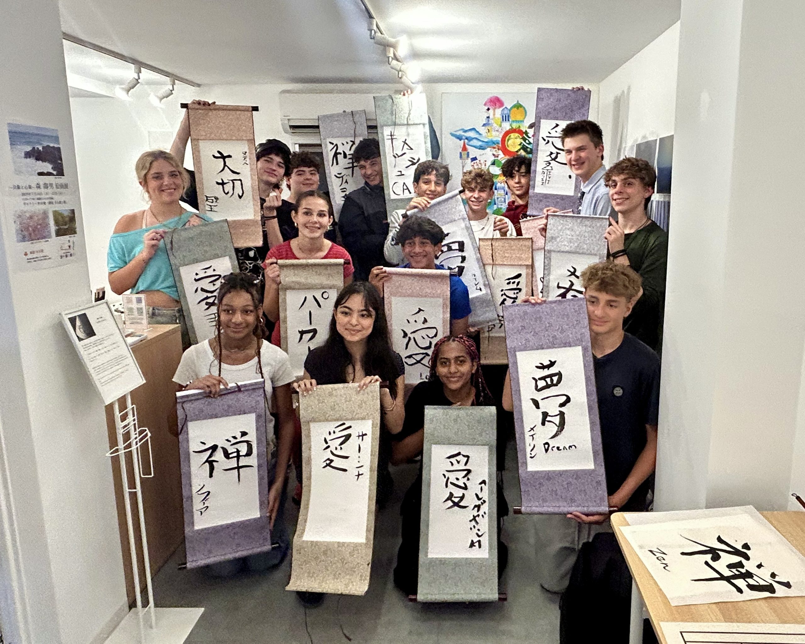 Teen travelers hold up mounted calligraphy scrolls with large brushed Japanese characters in a small gallery.