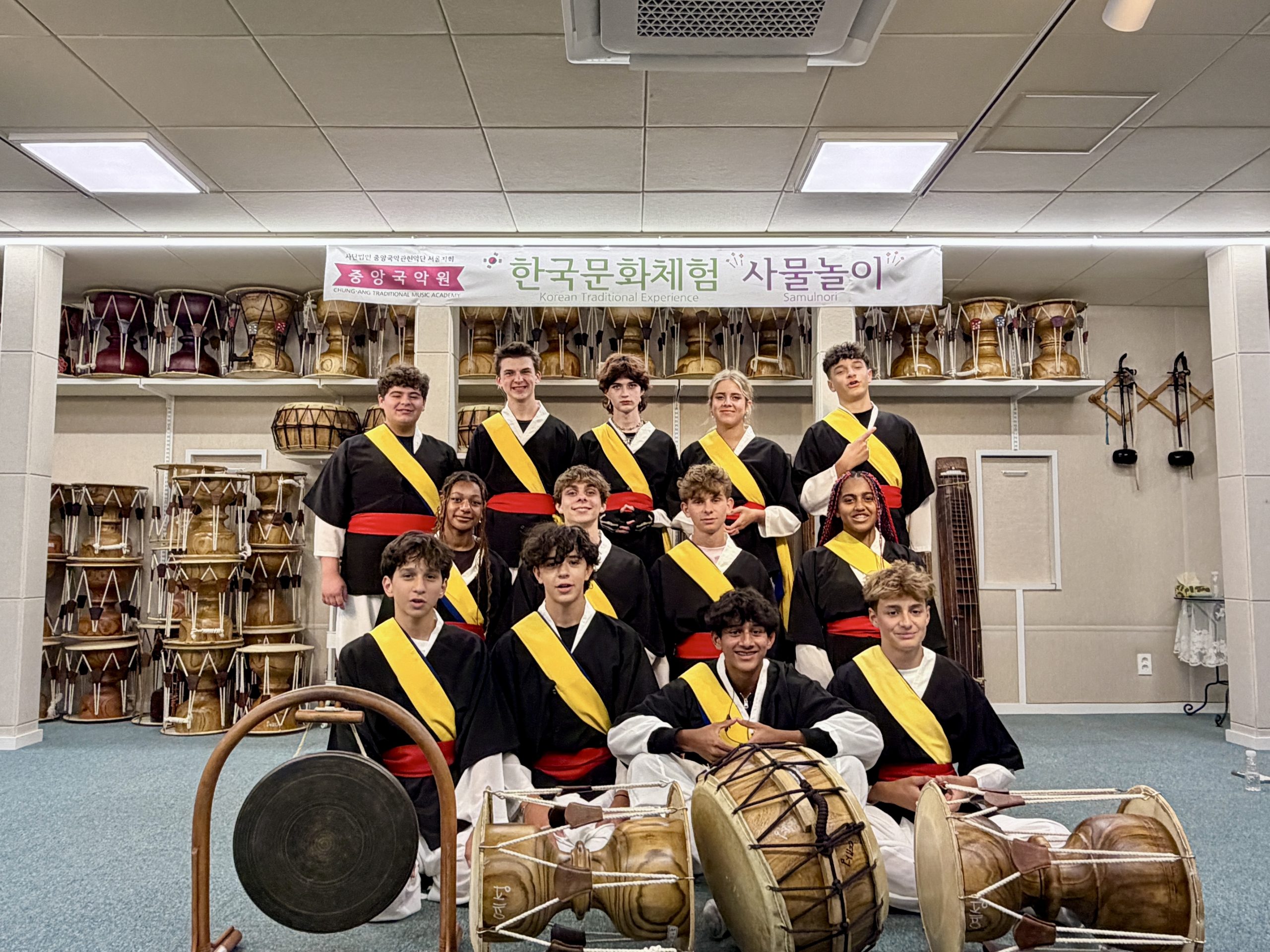 Teen travelers in black robes with yellow and red sashes pose with Korean drums and a gong under a Korean banner.