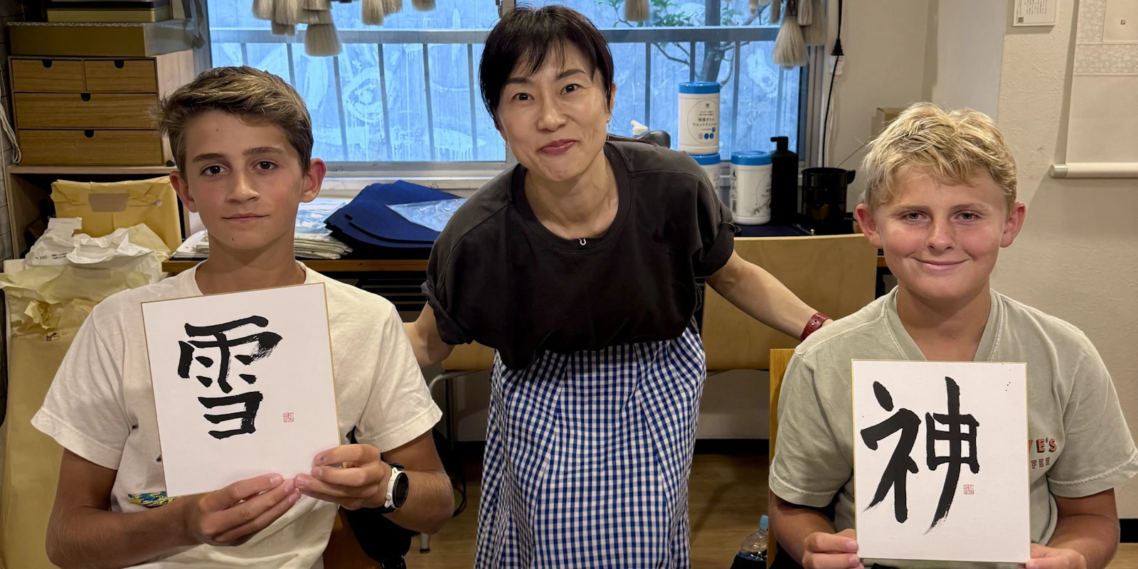 Two teen travelers hold up brushed kanji calligraphy boards beside their smiling teacher in a workshop.