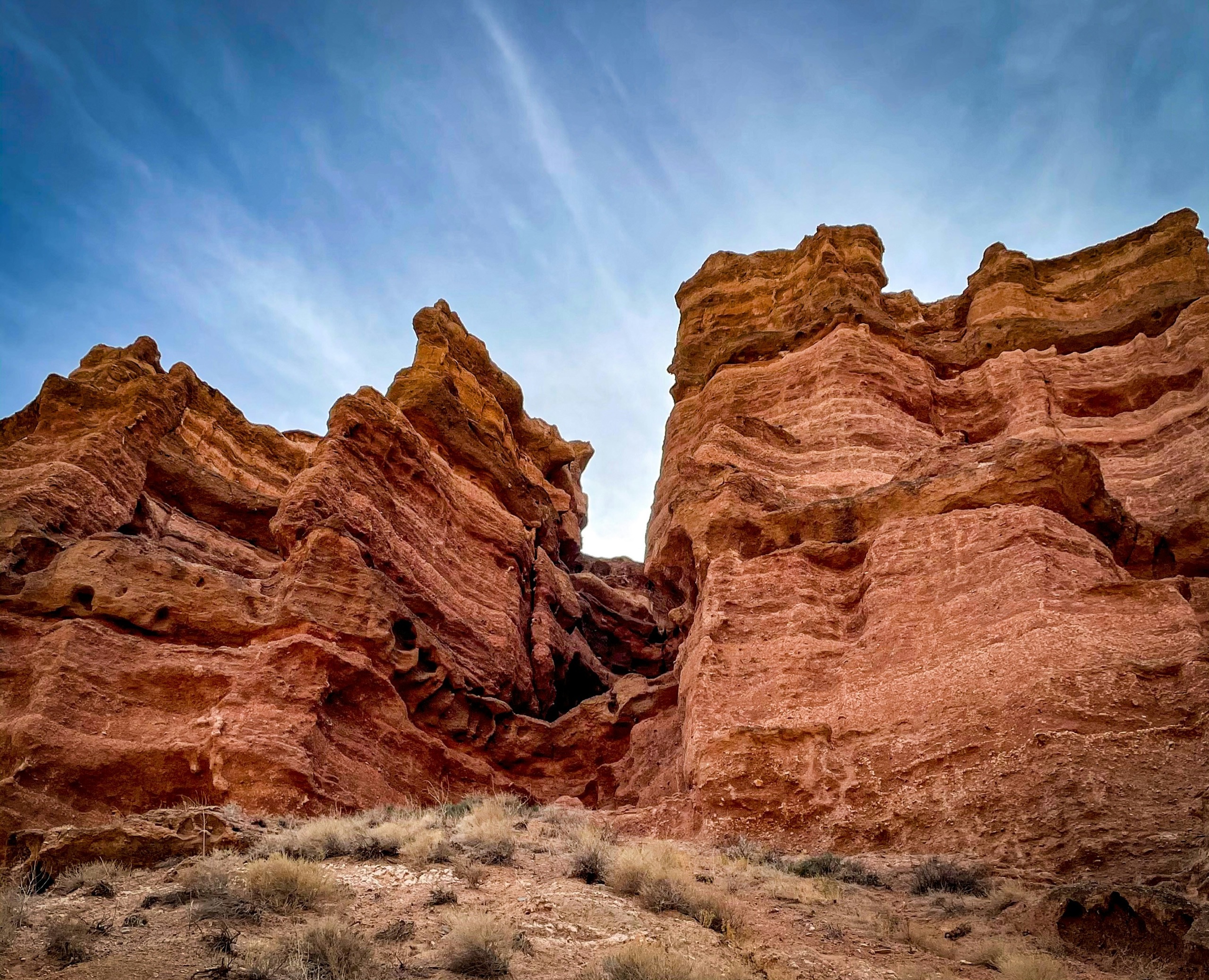 Red sandstone towers and ridges rise from scrub at the bottom of a canyon, their layered faces lit against a pale blue sky.