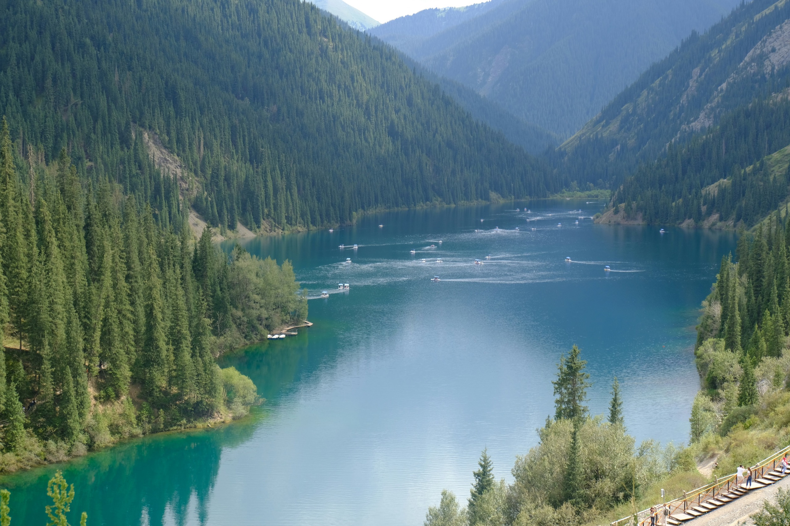 A long blue lake fills a steep valley of dark spruce forest, small boats dotted across the water and a boardwalk running along the near shore.