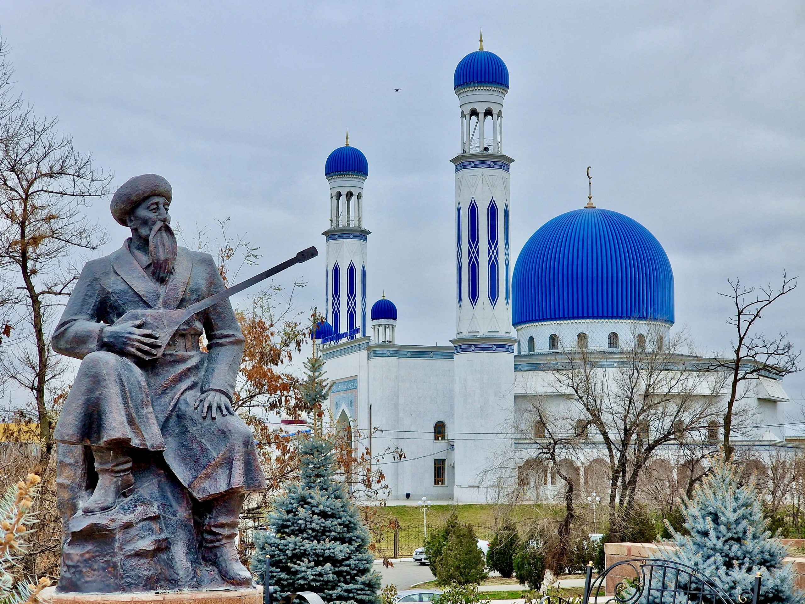 A bronze statue of a seated musician holding a dombra stands in front of a mosque with a blue dome and white minarets.