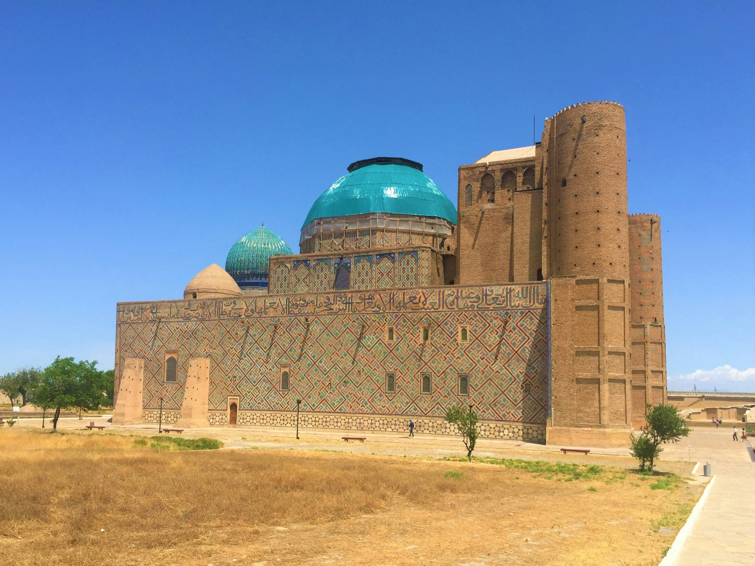 A vast brick mausoleum with turquoise domes and a wall of patterned tilework rises from a dry lawn under a deep blue sky, two small figures walking along its base.