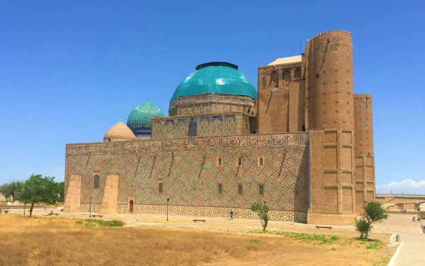 A vast brick mausoleum with turquoise domes and a wall of patterned tilework rises from a dry lawn under a deep blue sky, two small figures walking along its base.