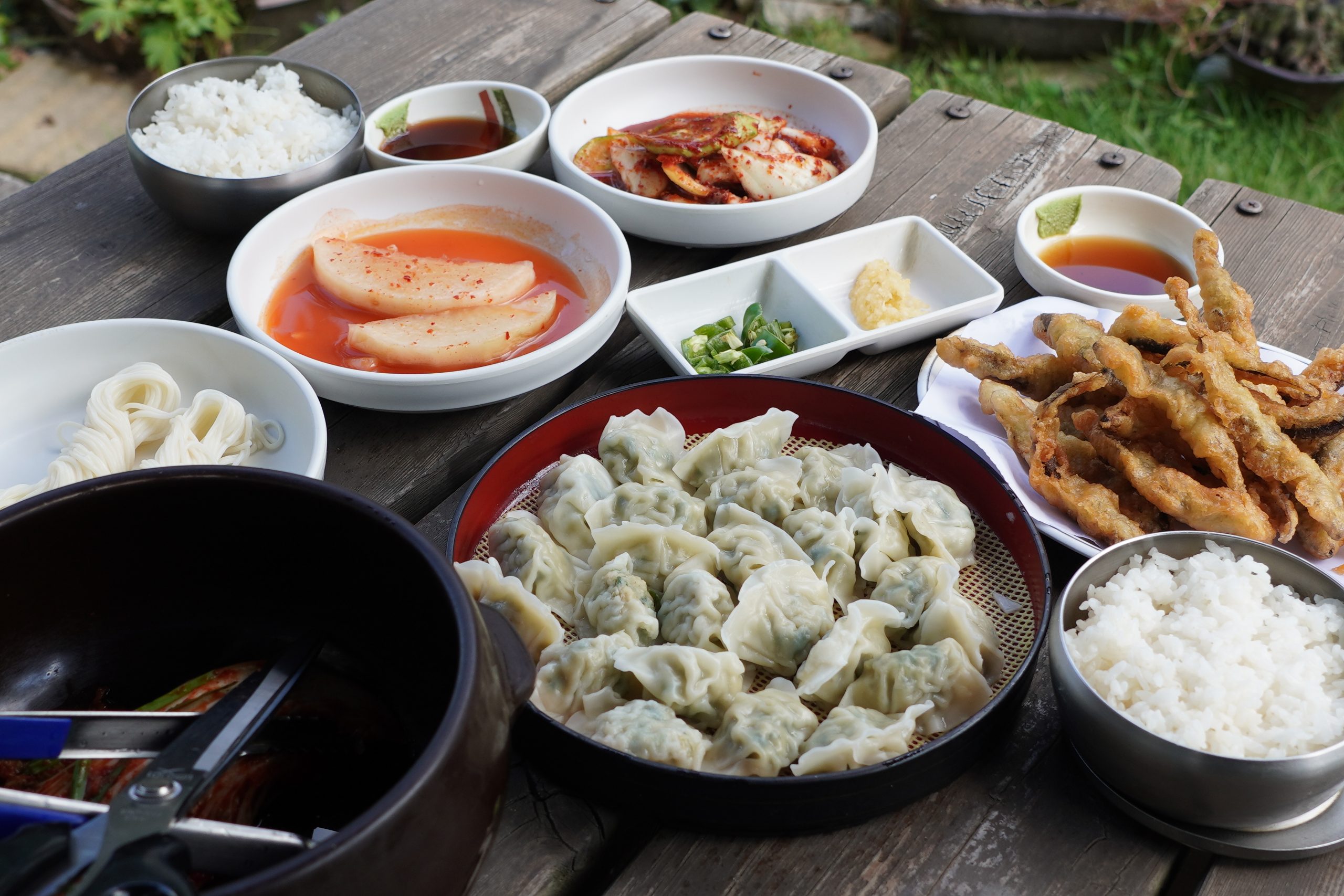A wooden table spread with Korean dumplings, kimchi, pickled radish, fried skewers, and rice bowls