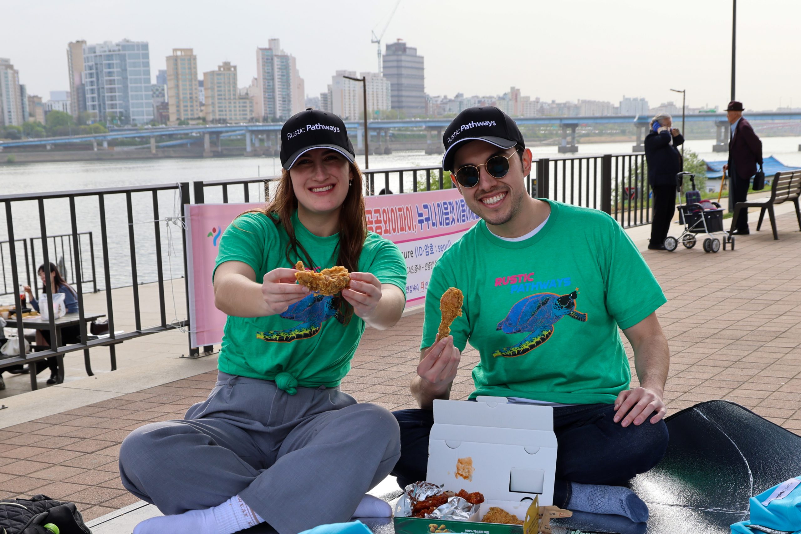 Students hold up fried chicken pieces while sitting along a riverside promenade with the city skyline behind them