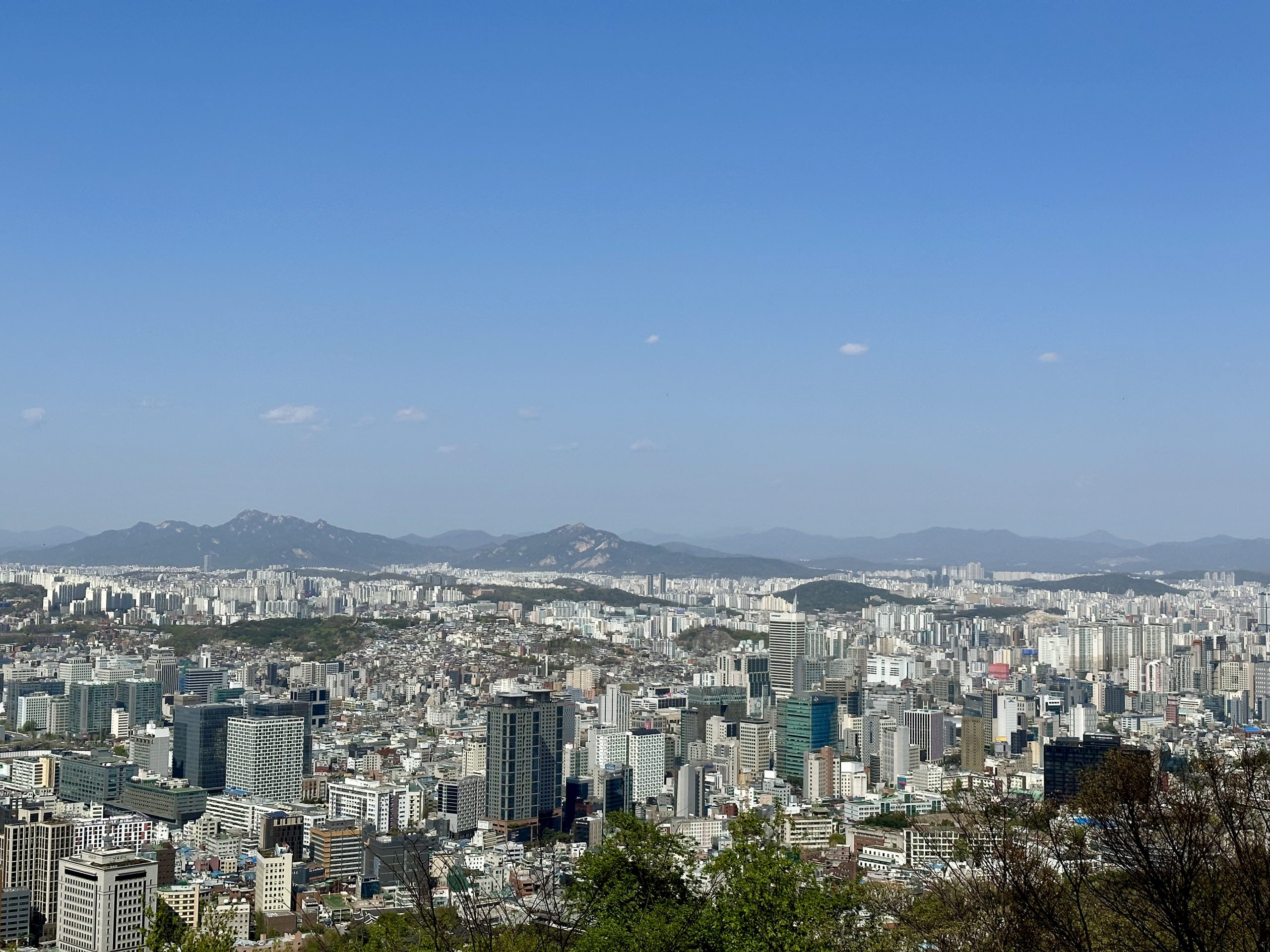 Skyline of Seoul viewed from Namsan Mountain, showing a mix of modern skyscrapers and surrounding mountain ranges under a clear sky.