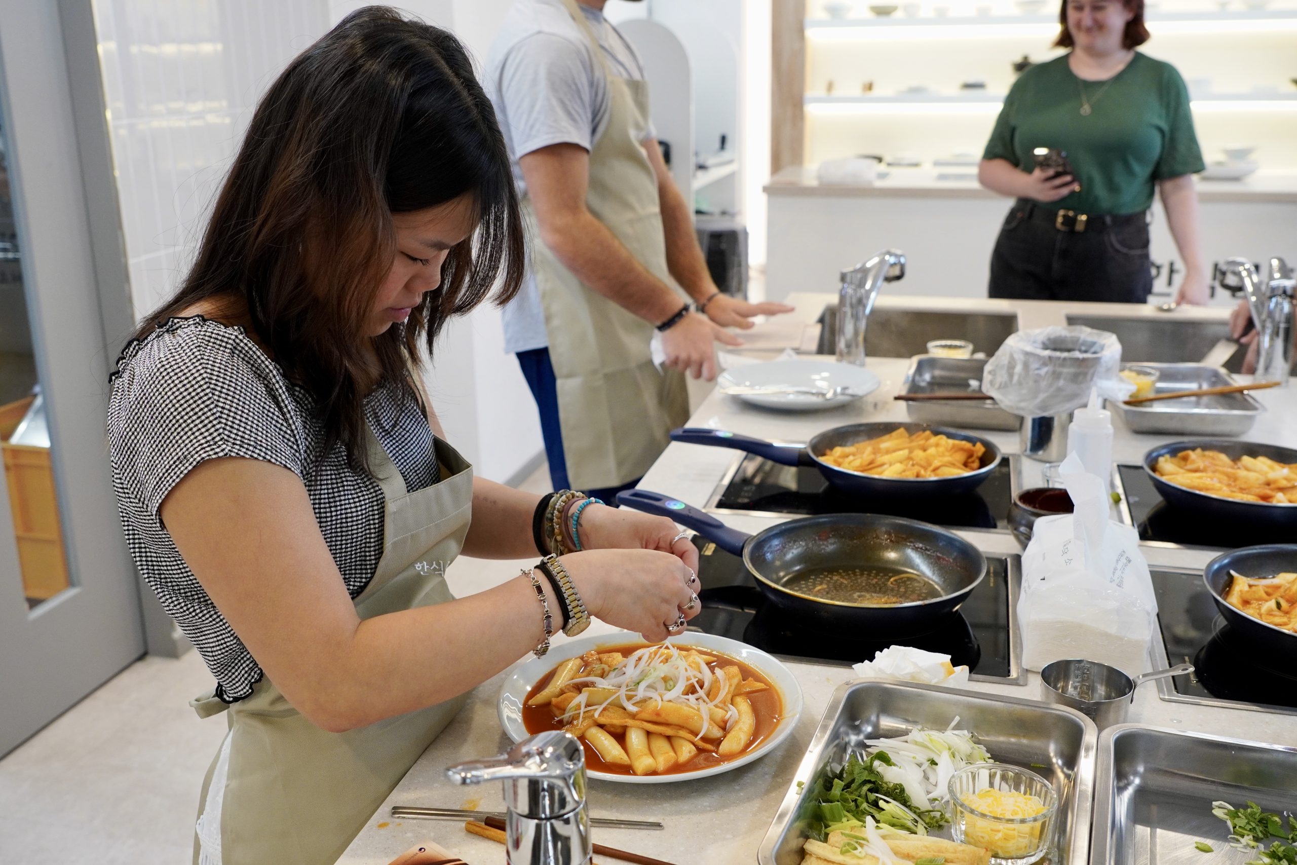 Student garnishes a plate of freshly made tteokbokki during a Korean cooking class, surrounded by ingredients and cooking tools.