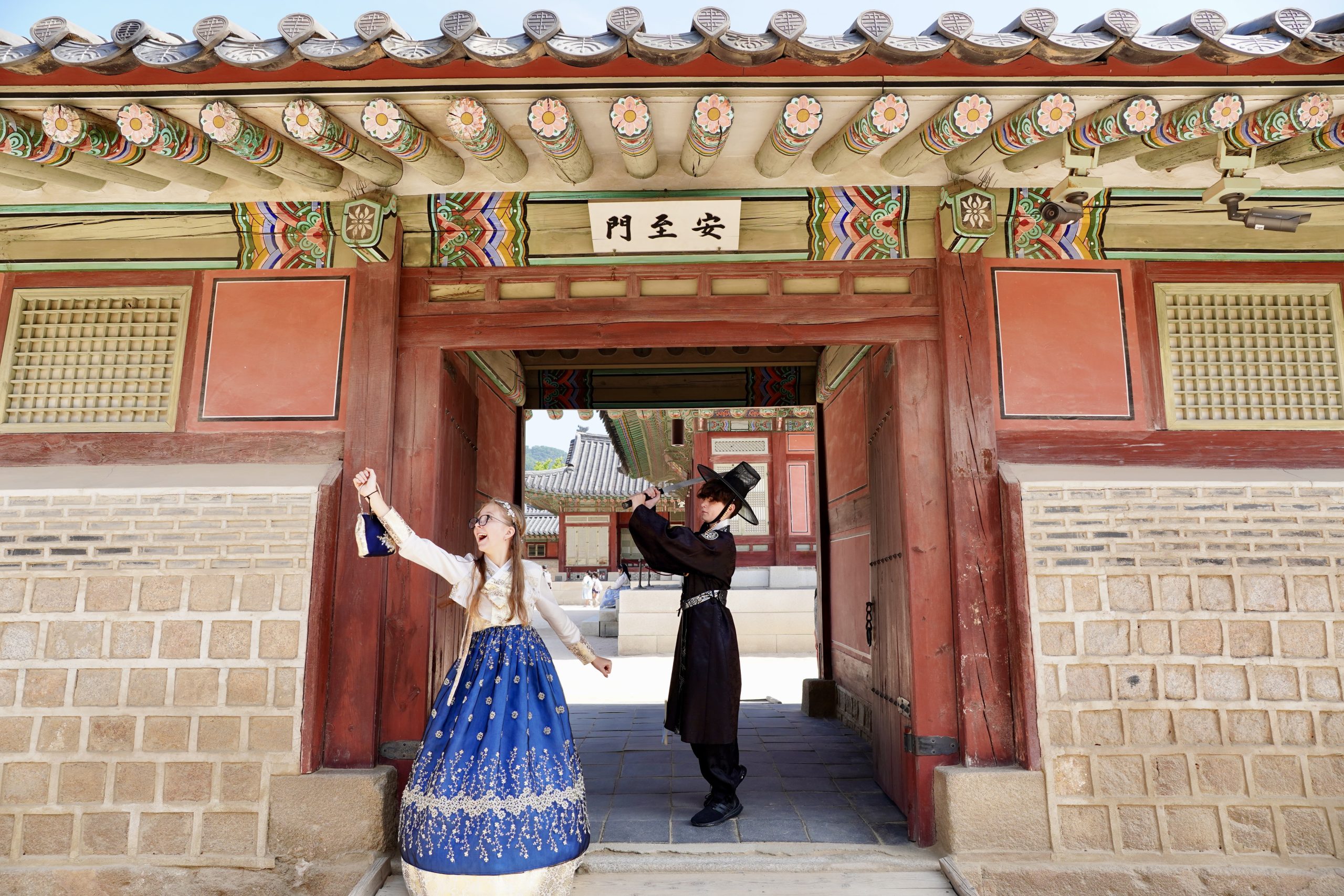 Two students wearing traditional hanbok pose joyfully at the entrance of a historic palace in Seoul, South Korea.