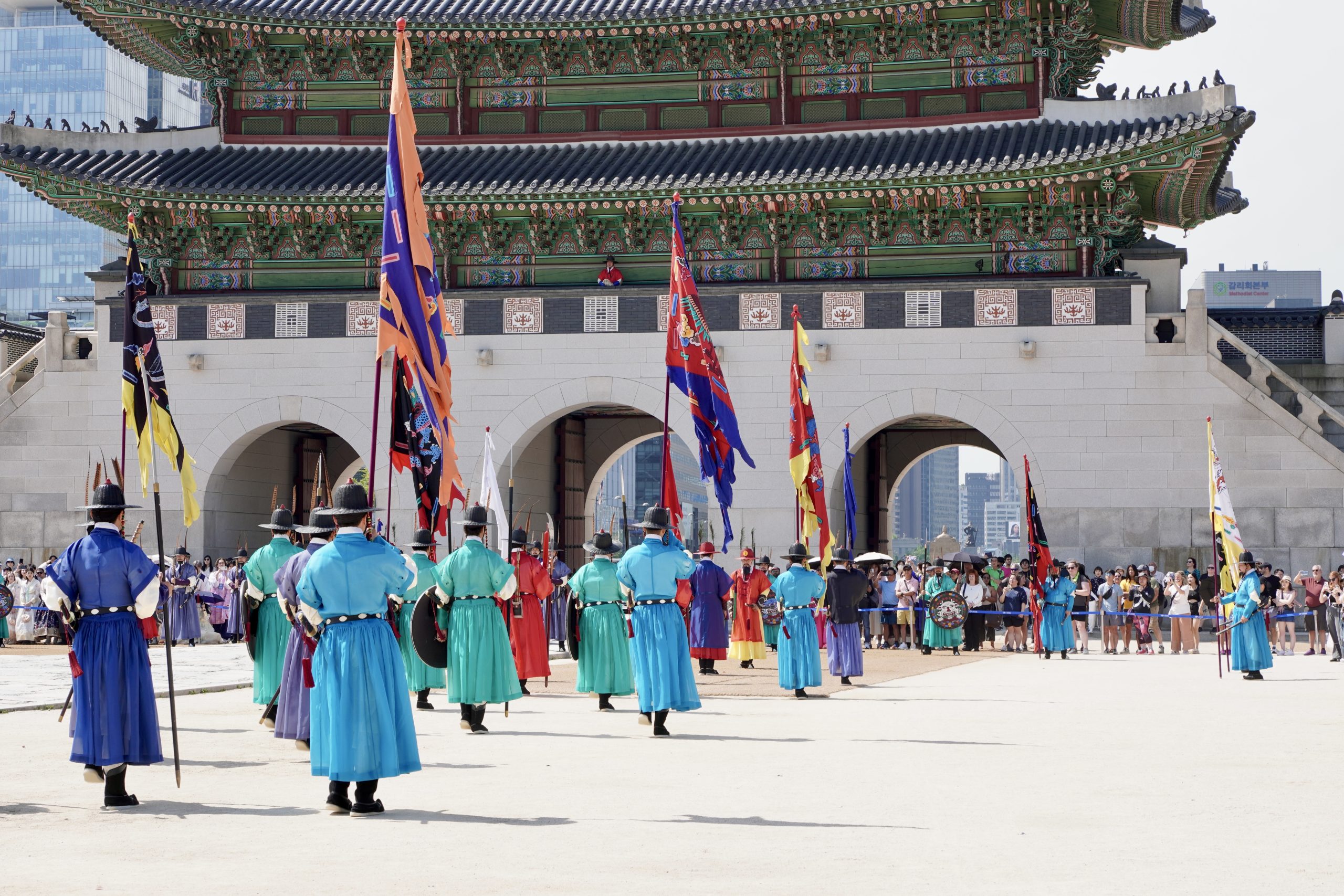 Changing of the guard ceremony in front of Gwanghwamun Gate at Gyeongbokgung Palace, with soldiers in colorful uniforms and a crowd of onlookers.