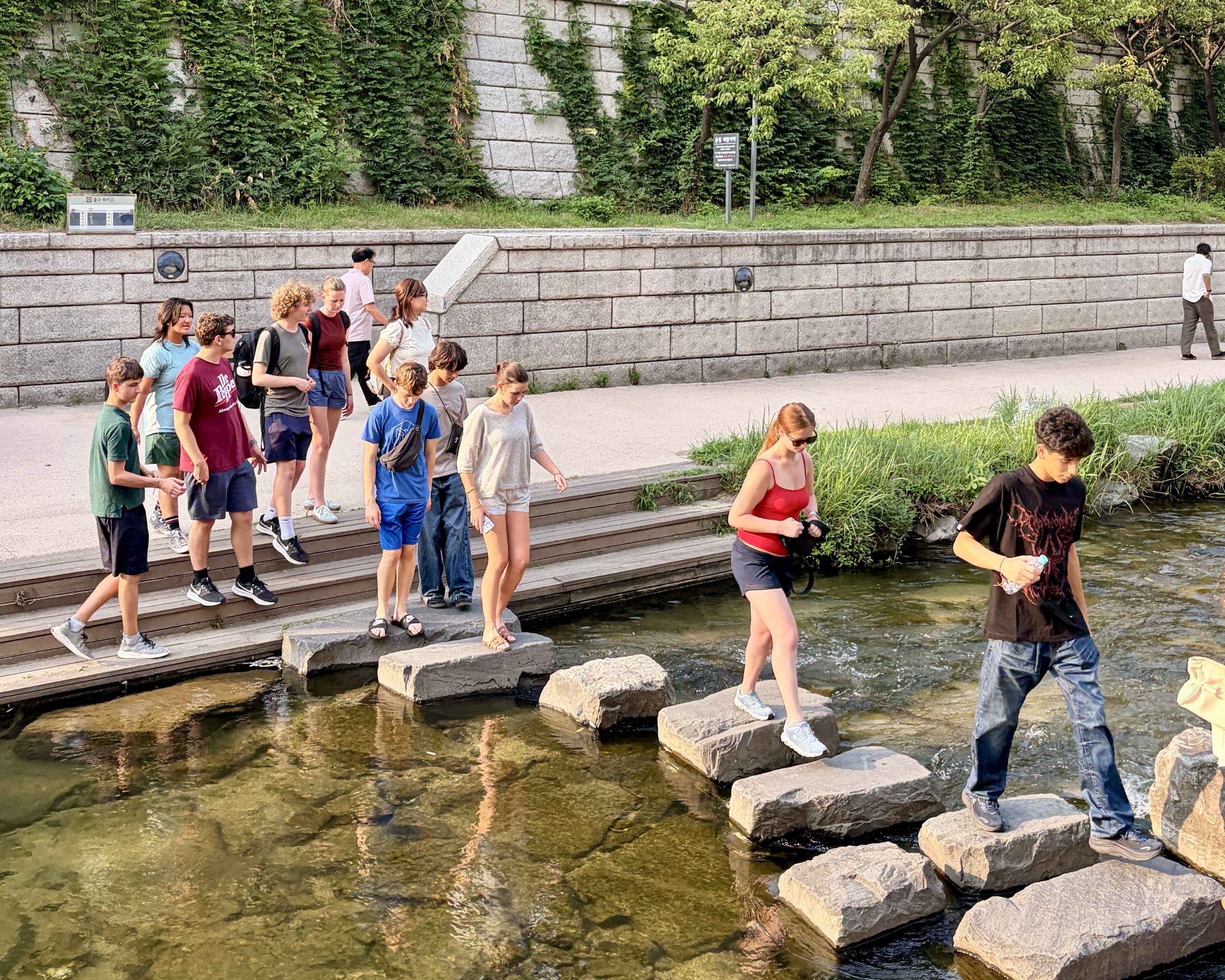 Students cross a line of stepping stones over a stream beside a palace wall