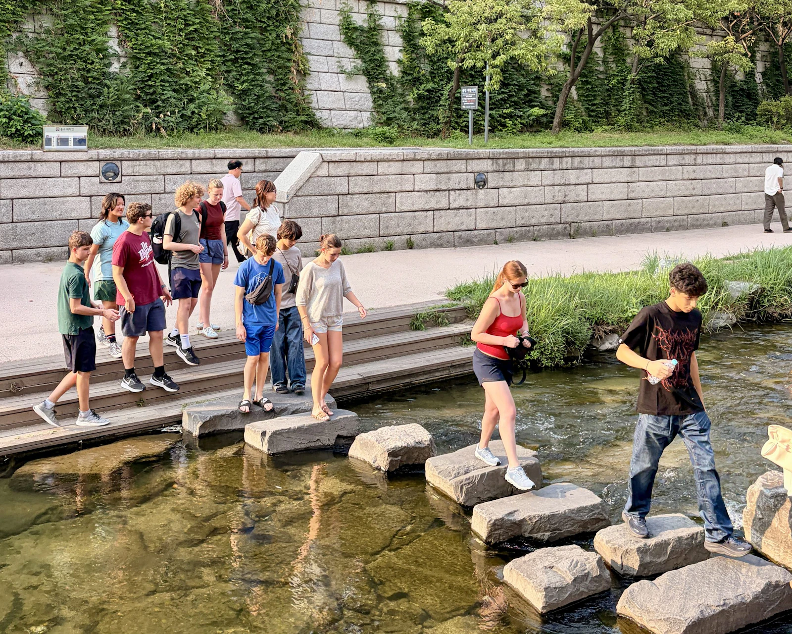 Students cross stepping stones over a shallow stream below a stone wall in Seoul