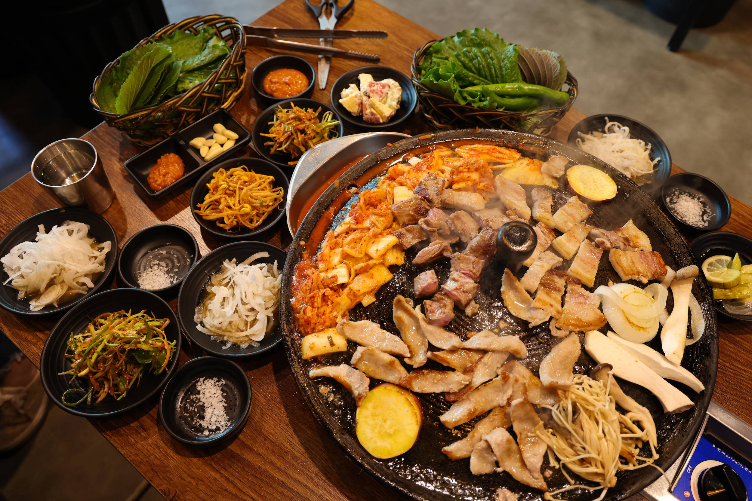 A Korean barbecue spread with grilled pork on a tabletop grill surrounded by banchan side dishes