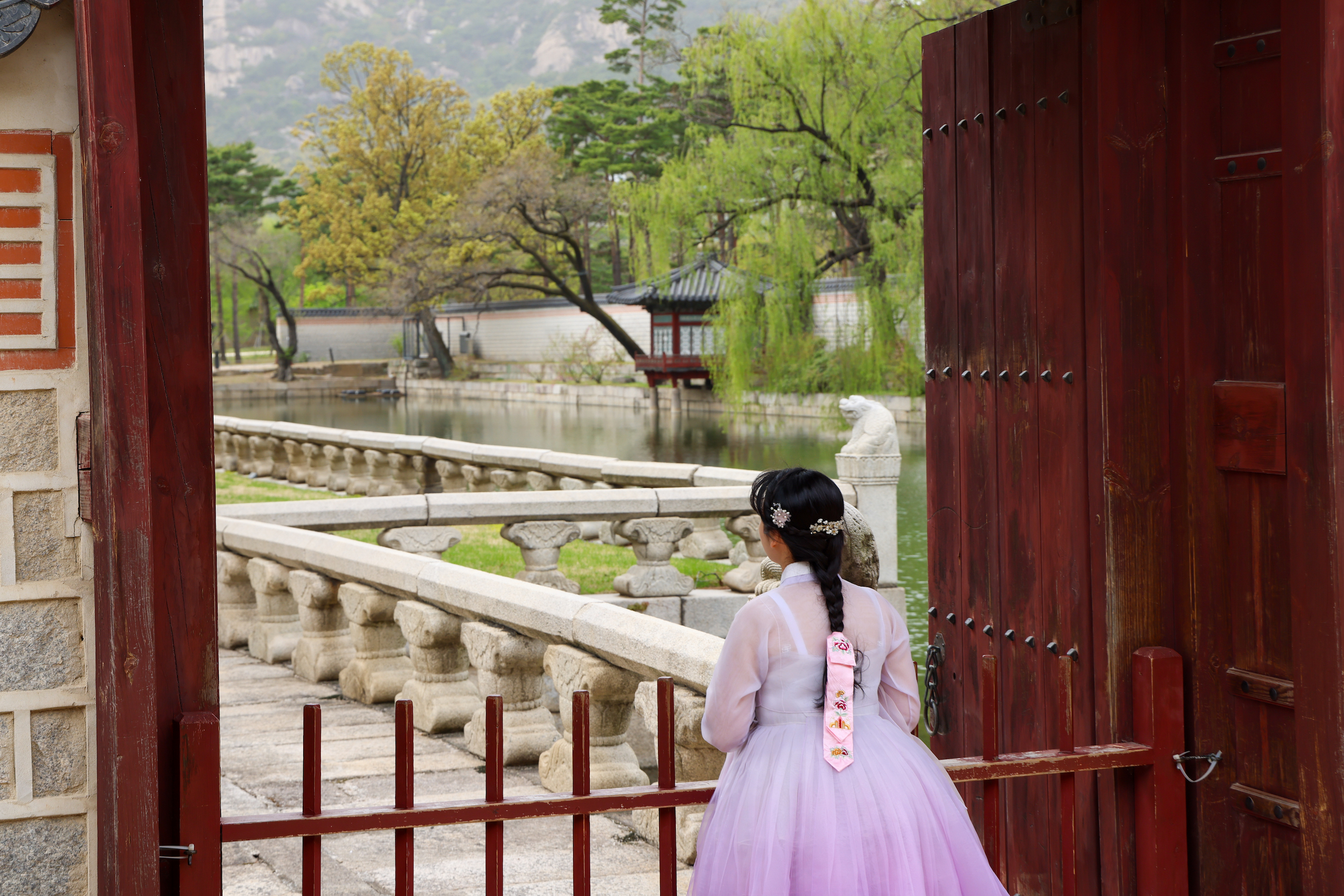Teens in traditional hanbok walk through a palace gate toward a stone bridge and pond