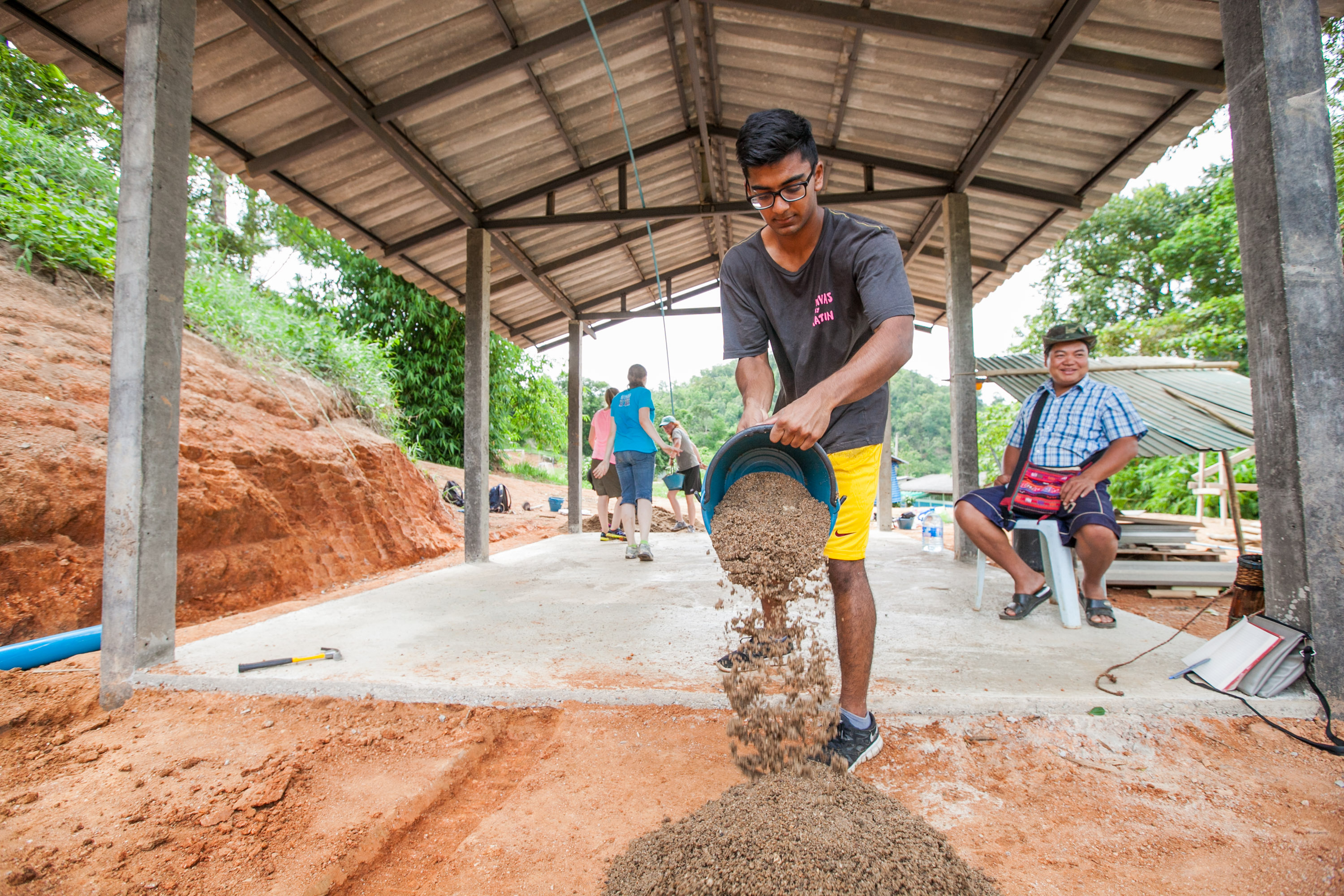 A student pours a wheelbarrow of gravel onto wet concrete at an outdoor construction site