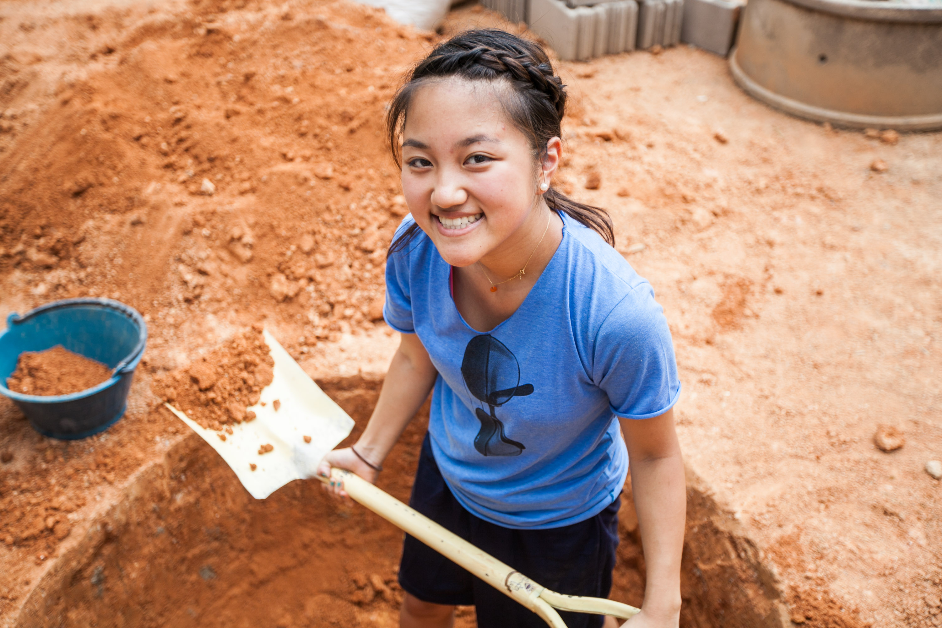 Students smile while digging with shovels at a red-dirt construction site