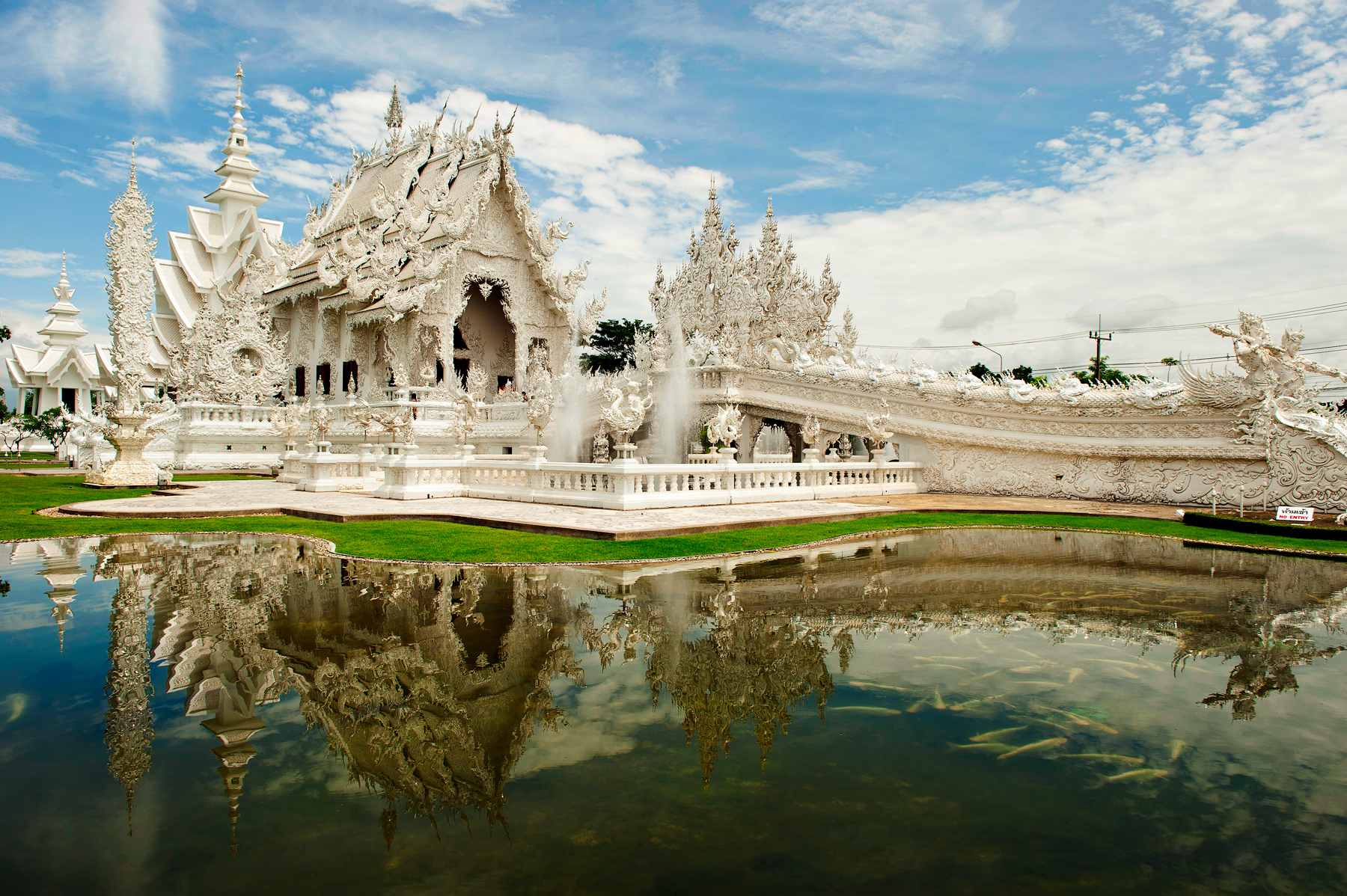 Ornate white temple in Laos with reflecting pool and fish beneath a blue sky