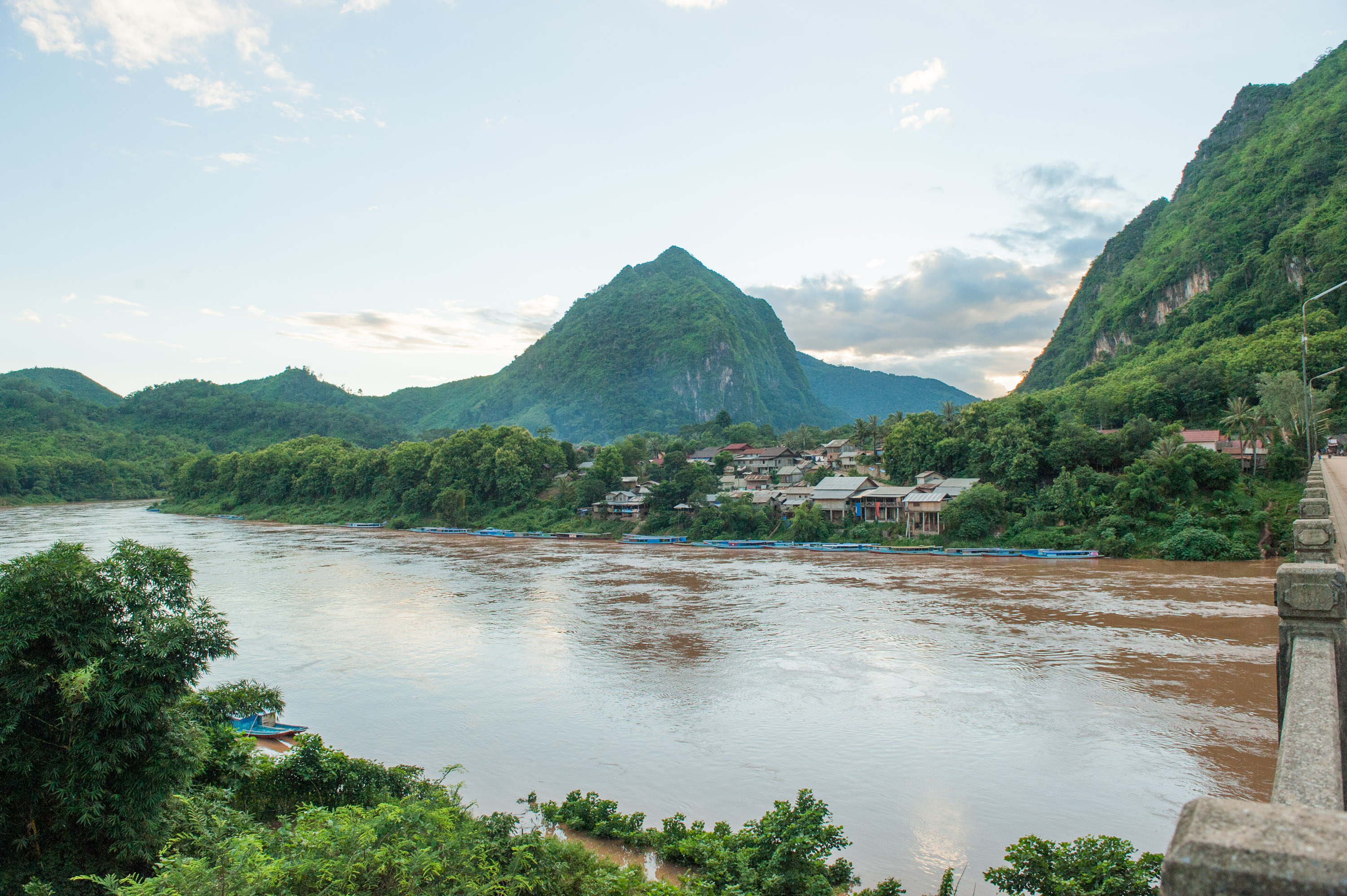 A sharp limestone peak rises above a riverside village, with longtail boats moored along the riverbank.