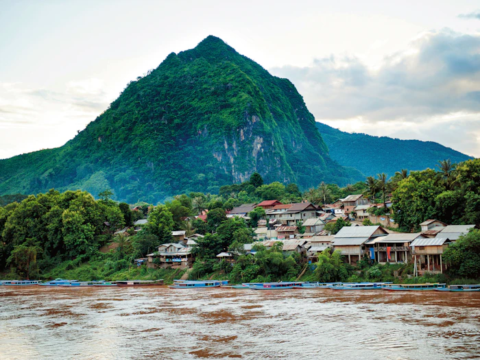 A sharp limestone peak rises above a riverside village, with longtail boats moored along the muddy riverbank.