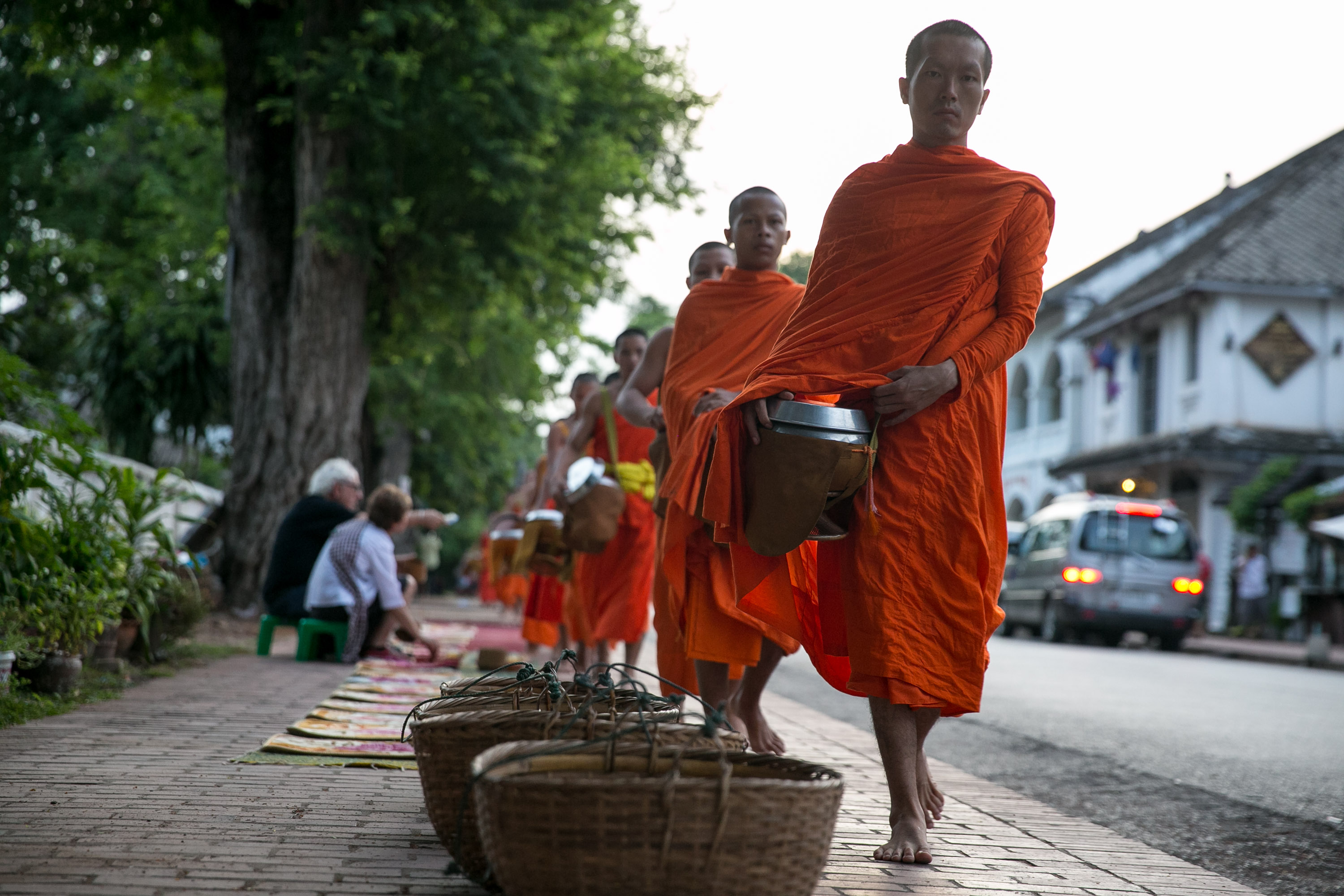Buddhist monks in orange robes walk along a street collecting morning alms