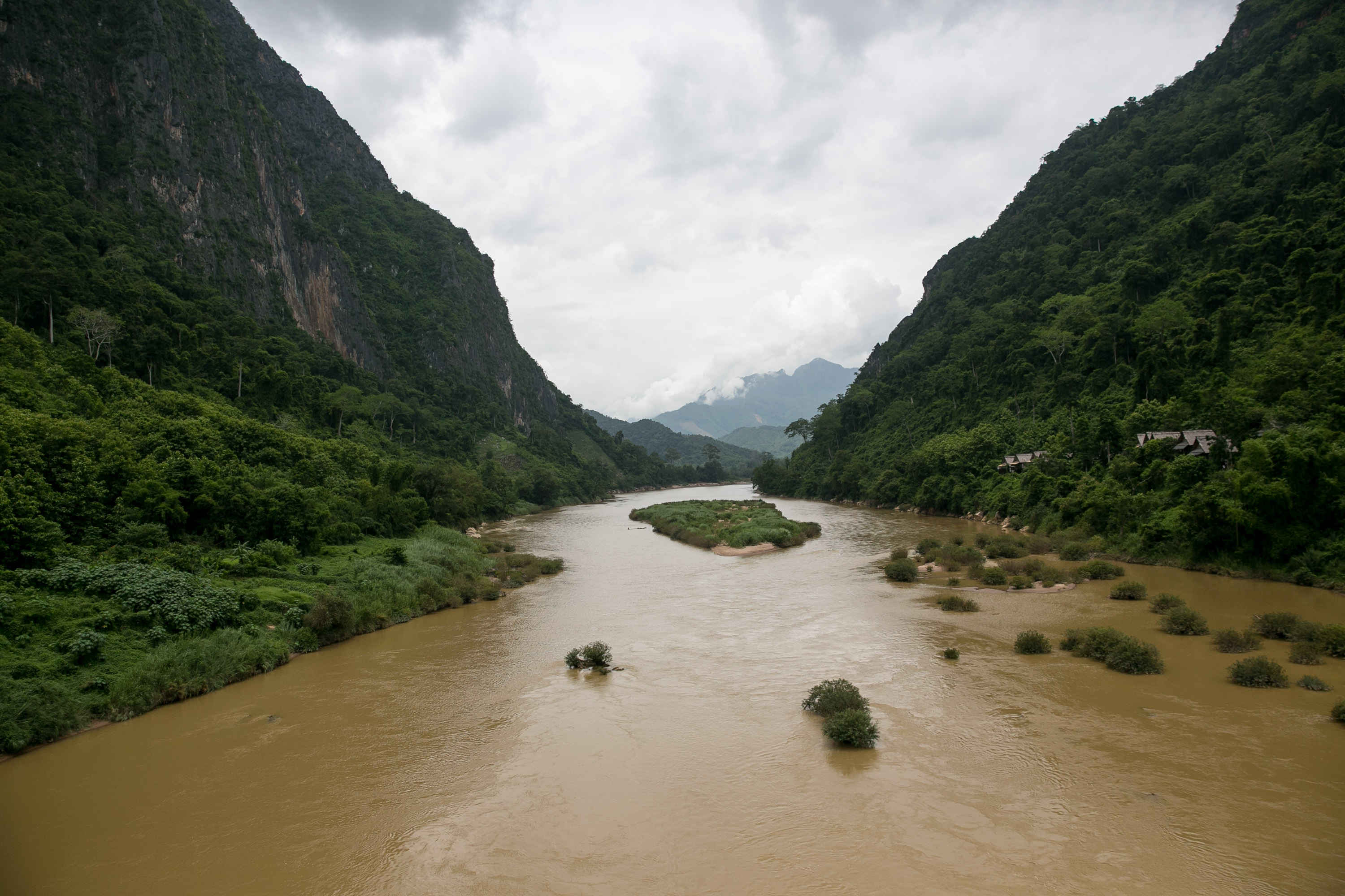 A muddy river in Laos winds between steep, jungle-covered mountains under cloudy skies