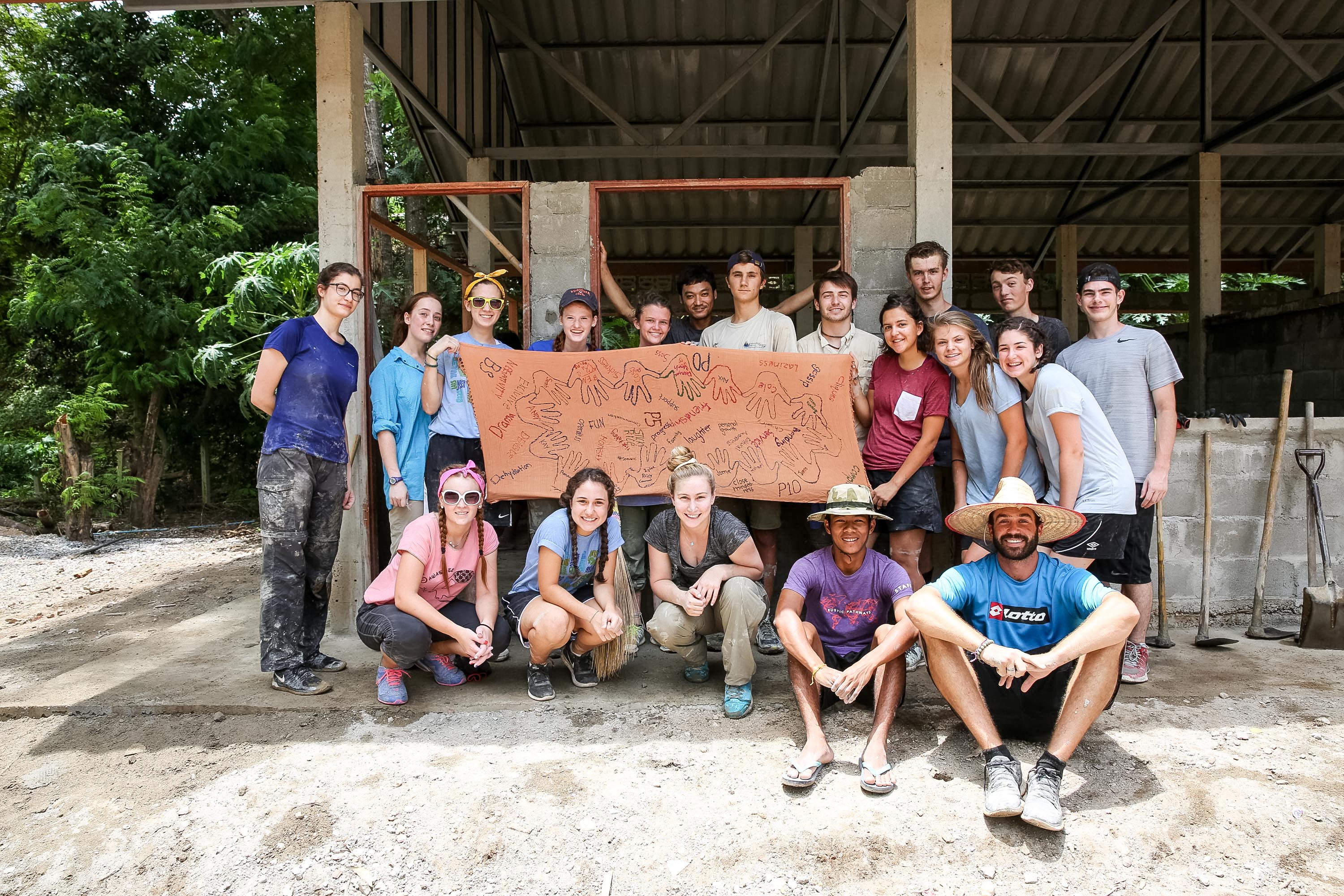 Students and leaders pose together under an open-sided shelter, some holding a painted banner