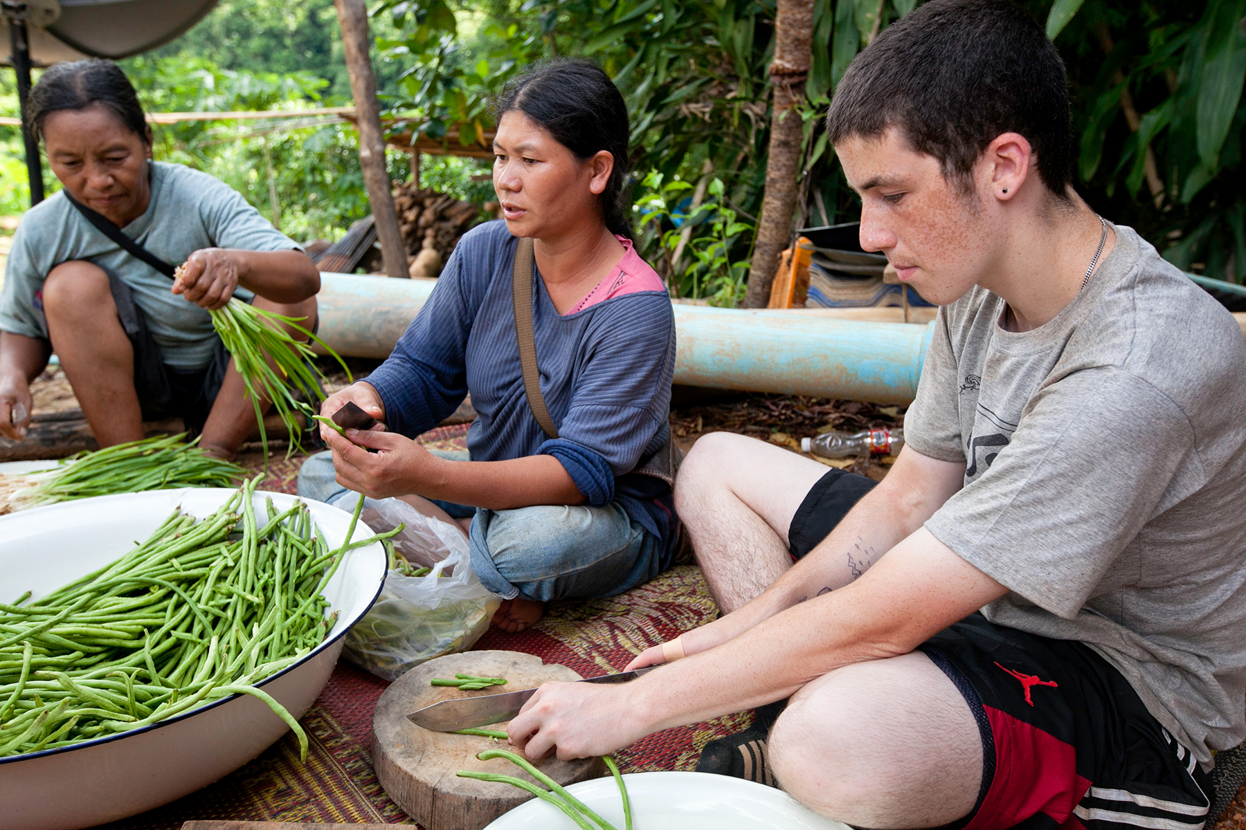 A school trip stops as a student joins local women preparing green beans on a woven mat