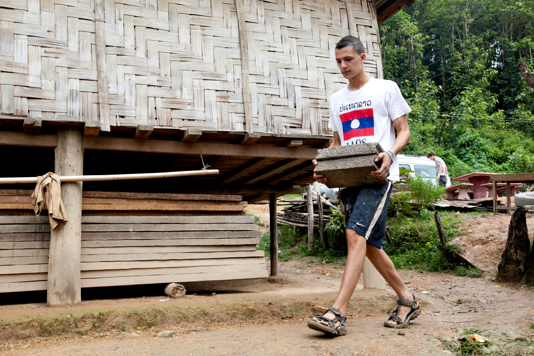 A student carries concrete blocks past a bamboo building on a service project